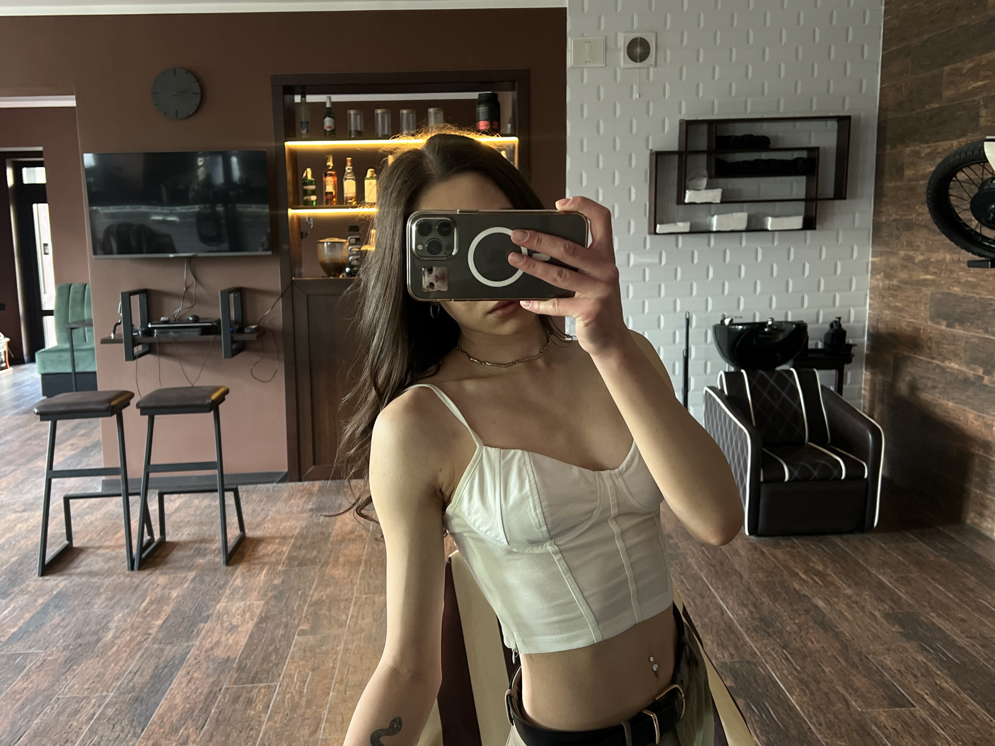 The image shows a woman holding a smartphone up to her face in a selfie pose inside what appears to be a salon or similar establishment. The woman has dark hair, a fair complexion, and is wearing a white top. She is partially obscuring her face with the phone. Her attire suggests a casual or relaxed setting.
The background reveals details of the salon interior. There are bar stools near a counter, and a television is mounted on the wall above. Behind her is a bar-like setup displaying bottles and glassware. There is a seating area with a unique chair design, and a wall with a textured pattern and wall decorations.
Given that the location is Ternopil, Ukraine, it is reasonable to assume that this is a beauty salon, hair salon, or a similar establishment in the city. The specific location or address is not evident from the image alone.