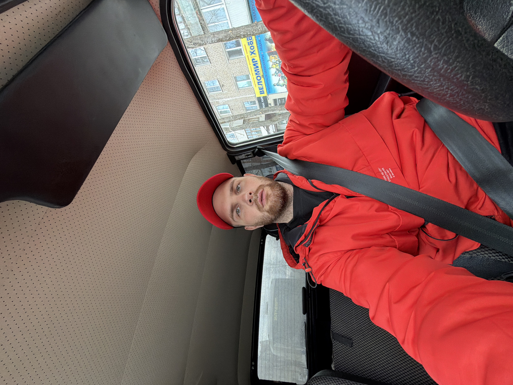 A selfie taken inside of a car. A fair-skinned man with a beard and wearing a red cap and red jacket is seen buckled into the driver's seat. The steering wheel of the car is visible in the upper right corner of the image. Through the car window, buildings can be seen with a yellow sign in Russian that translates to "Samara Velomir Hobby", and appears to be advertising bicycles. The seat of the car is a light tan color. The photo was taken in Chaikovsky, Russia.