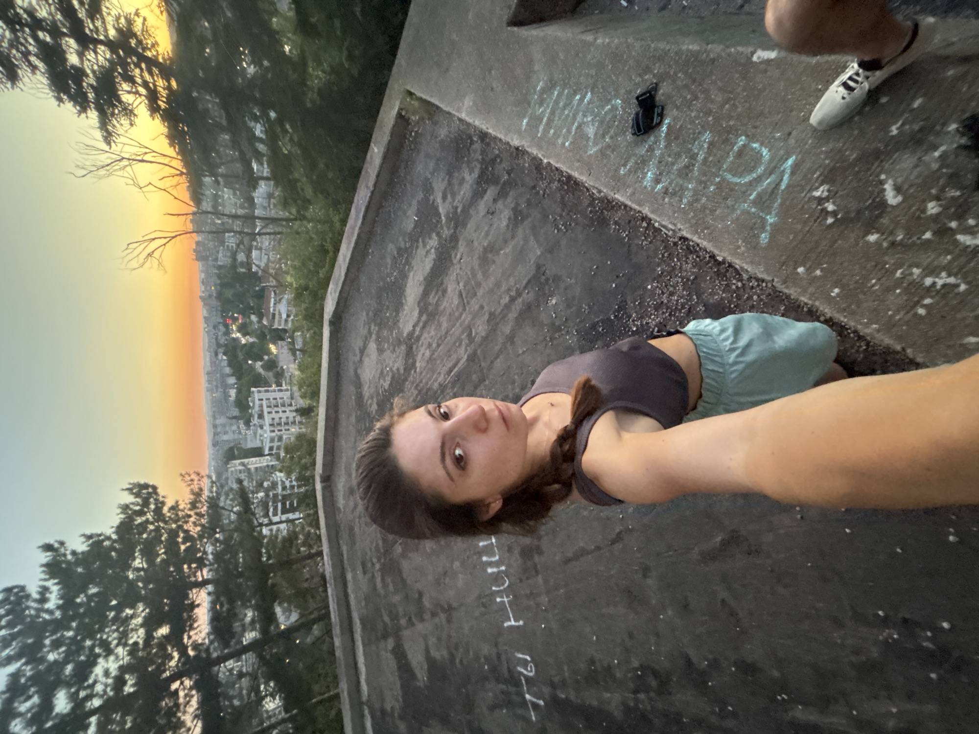A woman with brown hair in a braid takes a selfie on a concrete rooftop overlooking the city of Varna, Bulgaria, at sunset. She wears a dark gray tank top and light blue shorts. The sky displays a gradient of orange, yellow, and light blue. In the background, buildings and trees are visible. White chalk graffiti is present on the rooftop. A black object, possibly a camera accessory, and a white sneaker are on the edge of the rooftop. The overall scene suggests an urban landscape during a twilight hour.