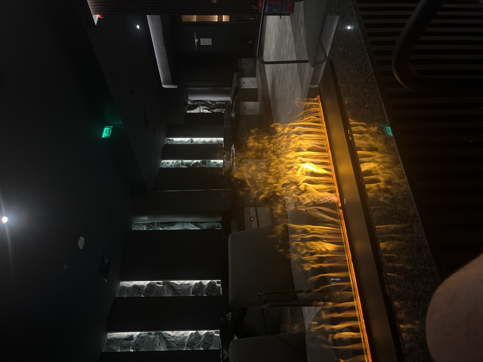 Here is a detailed description of the image:
**Scene:** The image depicts the interior of a dimly lit space, potentially a restaurant or bar. A prominent feature is a decorative "fire wall" with artificial flames. The room has modern architecture, incorporating dark tones and contrasting light elements. There are stairs with decorative panels underneath. Several chairs are visible. An exit sign is also visible on the wall.
**Objects:**
* Decorative "fire wall" with artificial flames
* Stairs with decorative panels
* Chairs
* Exit sign
**Location:** While the image does not provide explicit visual clues to confirm the location, the style and general aesthetic suggest a contemporary interior that could be found in many modern cities, including Sofia, Bulgaria.