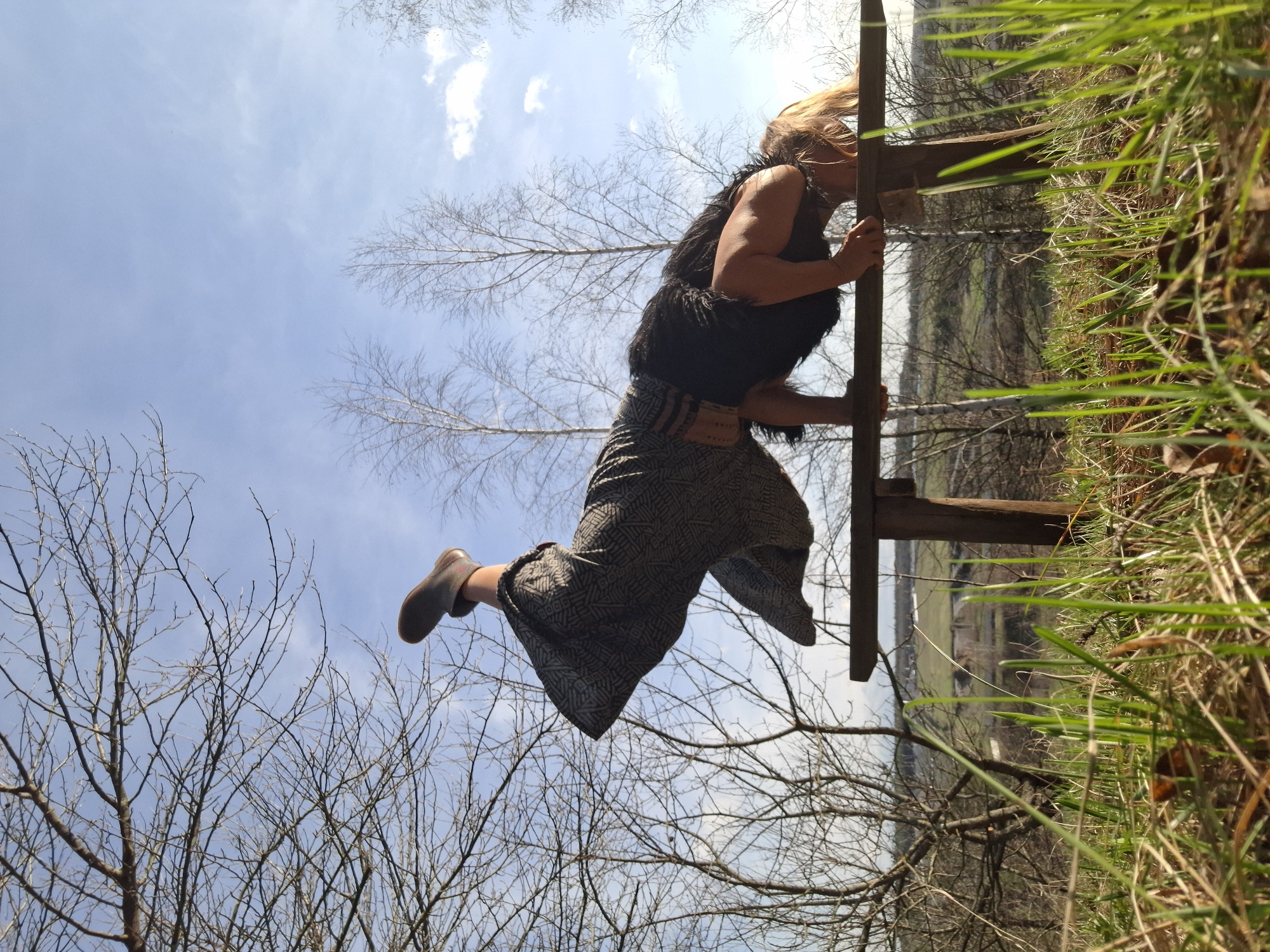 The image, rotated 90 degrees clockwise, captures a dynamic low-angle view of a person climbing or vaulting over a rustic wooden fence. The individual, with windblown blonde hair, wears a dark, possibly furry, sleeveless top or vest, along with loose-fitting patterned pants in shades of gray and black, and dark boots. Their hands firmly grip the top rail of the fence, while one leg is bent, suggesting mid-motion or a brief pause atop the barrier. A patterned belt with geometric designs is visible around their waist, but no discernible text can be read.
The foreground, framed by the right side of the rotated image, is dominated by lush, bright green grass and some dry foliage, indicating a vibrant natural setting. Behind the person and the fence, a canopy of bare deciduous trees with intricate branch networks fills the mid-ground, suggesting late winter or early spring. Above, a bright, clear blue sky with sparse white clouds completes the backdrop, illuminated by strong daylight. In the far distance, a rural landscape with fields or a valley can be faintly seen. The scene depicts an active moment in an outdoor, rural environment in Khlevnoye, Russia.
