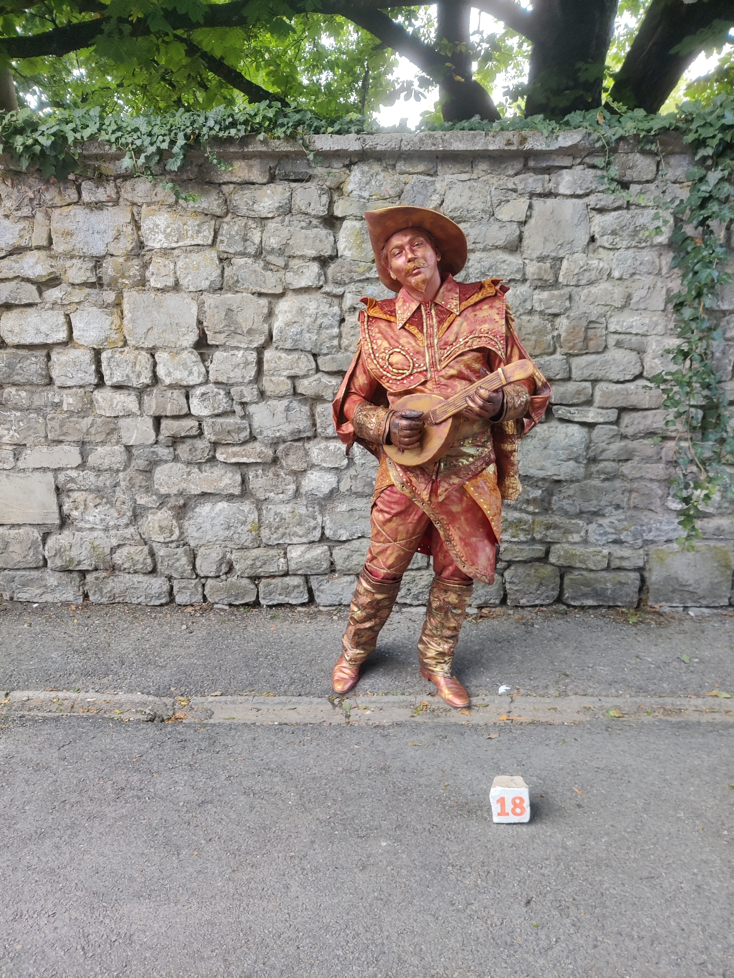 A costumed street performer, painted gold, stands in front of a stone wall in Marche-en-Famenne, Belgium. The performer wears a wide-brimmed hat and an elaborate gold and red suit, playing a lute-like instrument. A white cube with the number '18' in orange is placed on the asphalt in the foreground. Ivy climbs the stone wall behind the performer, and trees are visible overhead.