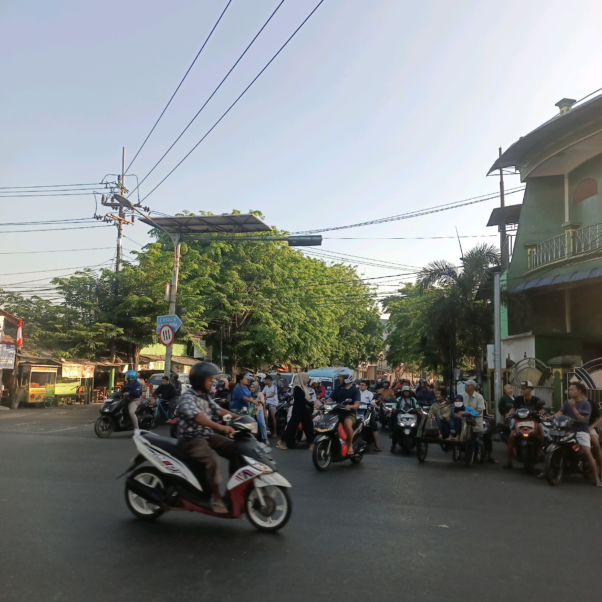 This image captures a bustling street scene in Surabaya, Indonesia, likely at an intersection, with a significant number of motorcycles and people. The time of day appears to be late afternoon or early evening, indicated by the warm, soft lighting and long shadows.
The foreground is dominated by the asphalt road, with several motorcycles in motion. A prominent motorcycle in the lower left is red and white, with a rider wearing a patterned shirt and helmet, captured in motion blur. To its right, another motorcycle is black, with a rider wearing a blue shirt and helmet. Further back, a variety of other motorcycles and their riders are visible, many also in motion.
In the background, a diverse crowd of people is present, some on motorcycles, others walking or standing. There are numerous stalls and small shops lining the side of the road, suggesting a commercial or residential area. One stall in the middle left appears to be selling food, with a display of what looks like fried snacks.
The environment is characterized by lush green trees providing ample shade, with power lines and utility poles crisscrossing the sky. A prominent green building with a curved roof and ornate balconies is visible on the right side of the image. A blue directional sign with white text and arrows indicates directions to "RSUD PRATAMA" and also features a traffic symbol with three horizontal lines in a circle, likely a speed limit indicator. The sky is clear and pale blue. The overall impression is one of everyday life and activity in a busy urban setting in Surabaya.
