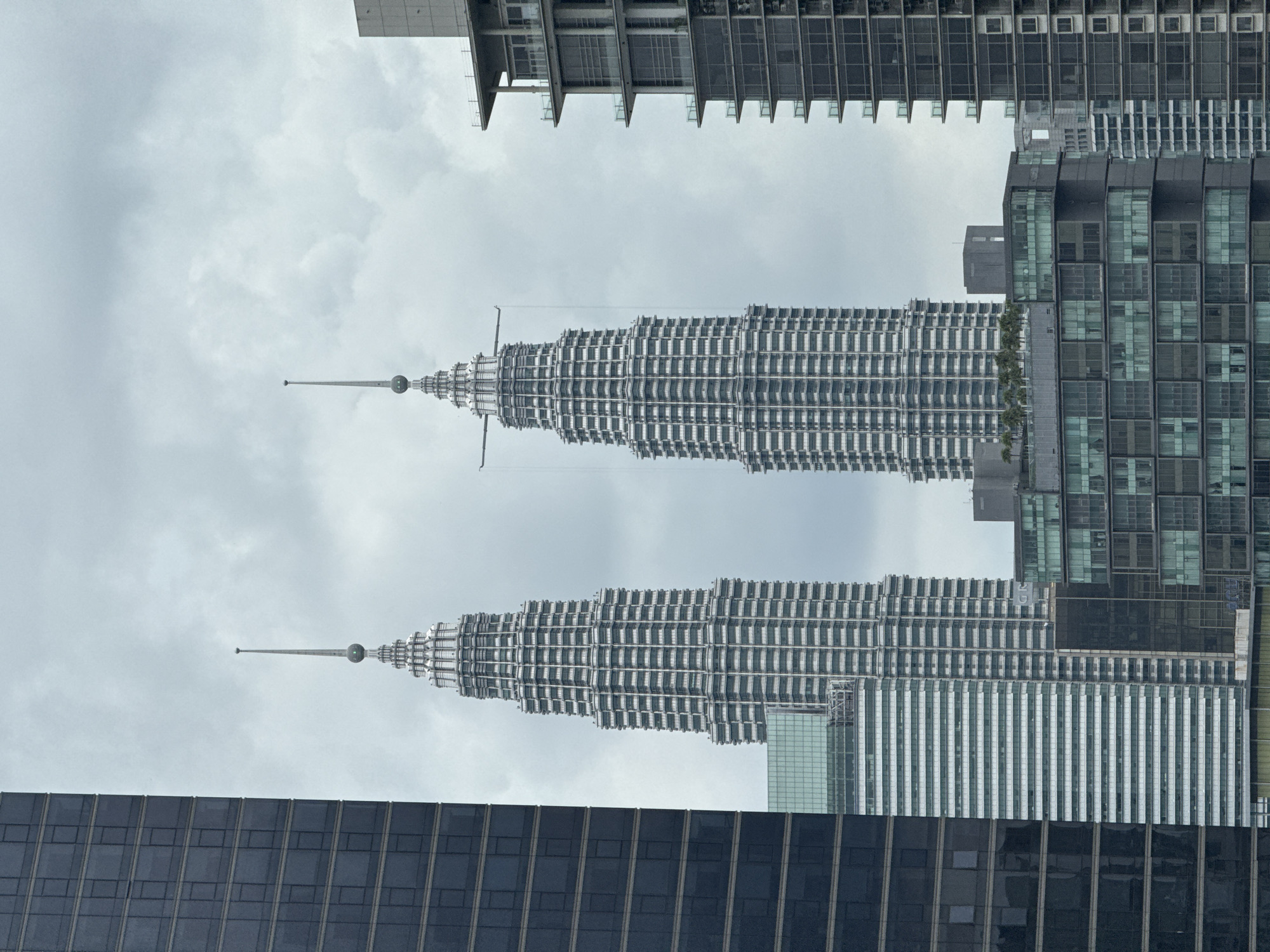The image shows the Petronas Twin Towers in Kuala Lumpur, Malaysia, framed by surrounding buildings. The towers, distinctive for their stacked, tapering design and spires, dominate the center of the frame against a cloudy sky. The lower half of the frame is filled with the dark glass and steel facade of a modern skyscraper, while the upper portion shows lighter-colored buildings, including those with horizontal, repeating architectural details. The towers are seen in their entirety, emphasizing their height and iconic silhouette within the urban landscape. No people are visible.