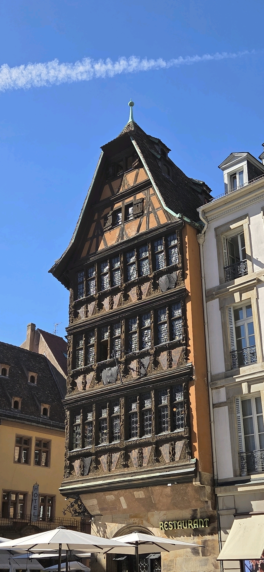 This is a vertical, eye-level shot of a narrow, ornate, timber-framed building in Strasbourg, France, with a bright blue sky and a faint contrail overhead. The building, identified as Maison Kammerzell, is a historic landmark located on the Place de la Cathédrale, adjacent to the Strasbourg Cathedral. The structure features multiple stories with intricately carved wooden details and numerous leaded glass windows. The roof is steep and covered in dark tiles, topped with a small green spire. To the right, a plainer, lighter-colored building with classical architectural elements is visible, its windows adorned with decorative ironwork. In the foreground, white outdoor umbrellas and tables suggest a restaurant or cafe setting. Another building, a hotel with a vertical "HOTEL" sign, is partially visible to the left. The image is well-lit, indicating it was taken during daylight hours. There are no people prominently featured in the image.