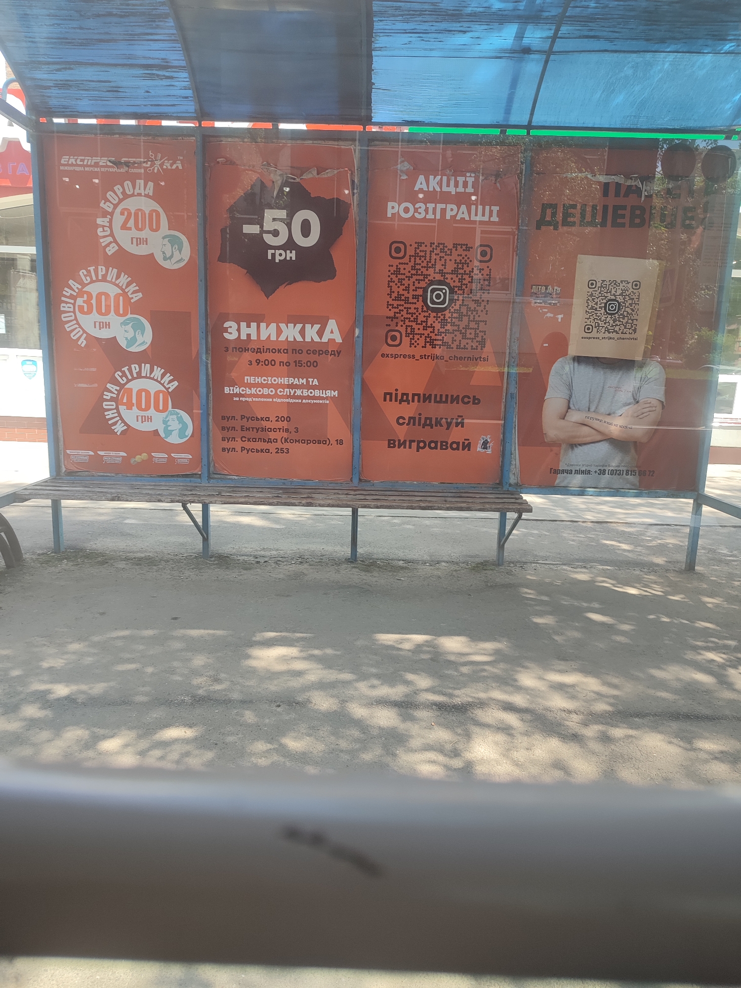 The image shows an outdoor bus stop shelter with advertisements on the back panels. The advertisements are in Ukrainian and promote discounts and contests for a barber shop named "Експрес Стрижка". One panel advertises a 200 UAH discount on a beard trim, a 300 UAH discount on a men's haircut, and a 400 UAH discount on a women's haircut. Another panel offers a 50 UAH discount from Monday to Wednesday between 9:00 AM and 3:00 PM, with additional discounts for pensioners and military personnel. There are QR codes on the panels, linking to the barber shop's Instagram page, and a phone number for a hotline is provided: +38 (073) 815 66 72. A person wearing a t-shirt and a paper bag with a QR code on their head stands in front of one of the advertisements. The bus stop is likely located in Chernivtsi, Ukraine, given the Ukrainian language of the advertisements and the mention of "Chernivtsi" in the Instagram handle "exspress_strijka_chernivtsi". The shelter has a blue translucent roof and a wooden bench. Sunlight casts shadows of tree branches on the ground.