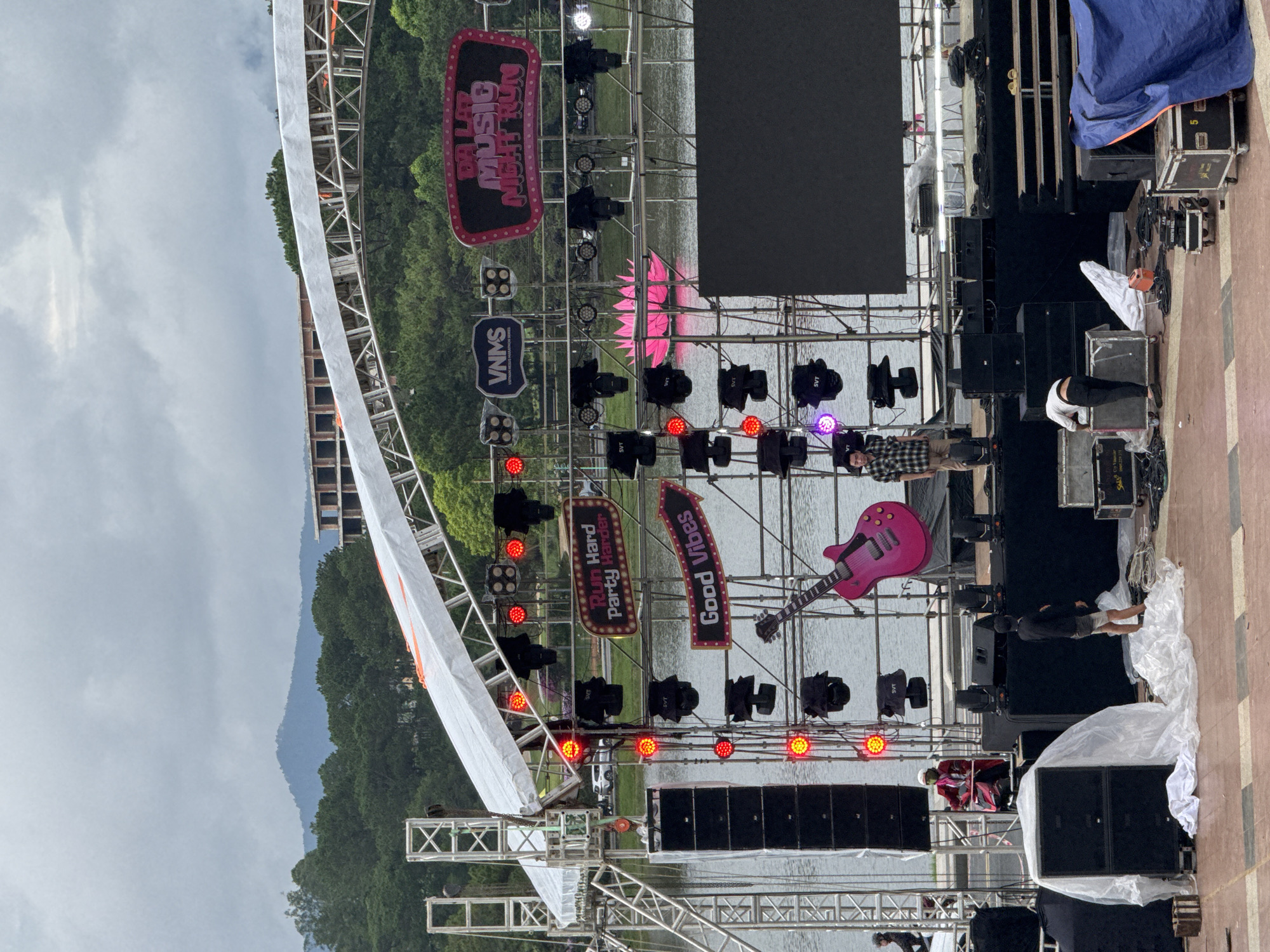 This outdoor scene captures the setup or preparation for a music event in Da Lat, Vietnam. A large stage structure, constructed from metal scaffolding, is positioned over or beside a body of water, likely a lake. In the background, lush green trees line the far shore, and distant mountains rise under a cloudy, overcast sky.
The stage is equipped with numerous speakers, stacked on platforms and around the base, and various stage lights, some emitting red, white, or purple glows, are mounted on the scaffolding. Two large screens are visible on the stage; one is a dark, blank rectangular screen, and another displays text. Decorative elements include a prominent pink electric guitar prop suspended near the stage, and a pink lotus flower-like sculpture floating in the water. Several signs are attached to the stage structure, reading "DA LAT MUSIC NIGHT RUN," "VNMS," "Run Hard Party Harder," and "Gooood Vibes."
Several individuals, presumably event staff or crew, are present on and around the stage. One person in a plaid shirt stands on the main stage platform, while another person in a dark top and light shorts is near equipment covered in white plastic sheeting on the paved foreground. Another individual is bending over near flight cases and speakers. Equipment, including speaker components labeled "SVT" and "JVC," is seen scattered around, some covered with blue tarps and white plastic. The overall impression is one of active preparation for an upcoming performance or festival, with no audience visible yet.