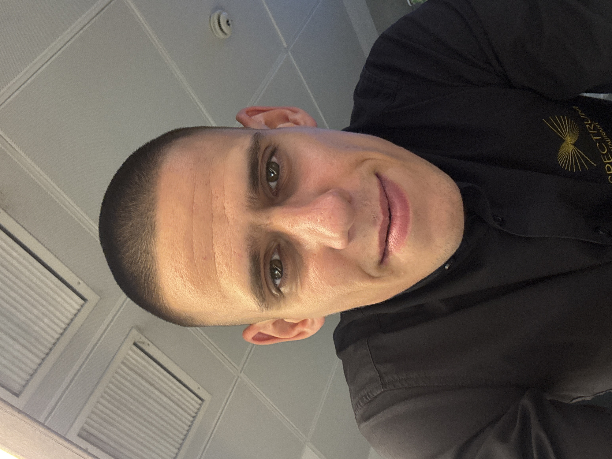 A man with a buzzed haircut and brown eyes is looking directly at the camera. He is wearing a black collared shirt with a logo on the left side that reads "SPECTRUM RESTAURANT". The man appears to be indoors, with a white ceiling with grid patterns and ventilation ducts visible above him. There is also a smoke detector on the ceiling. The background suggests an office or restaurant environment in Sofia, Bulgaria.