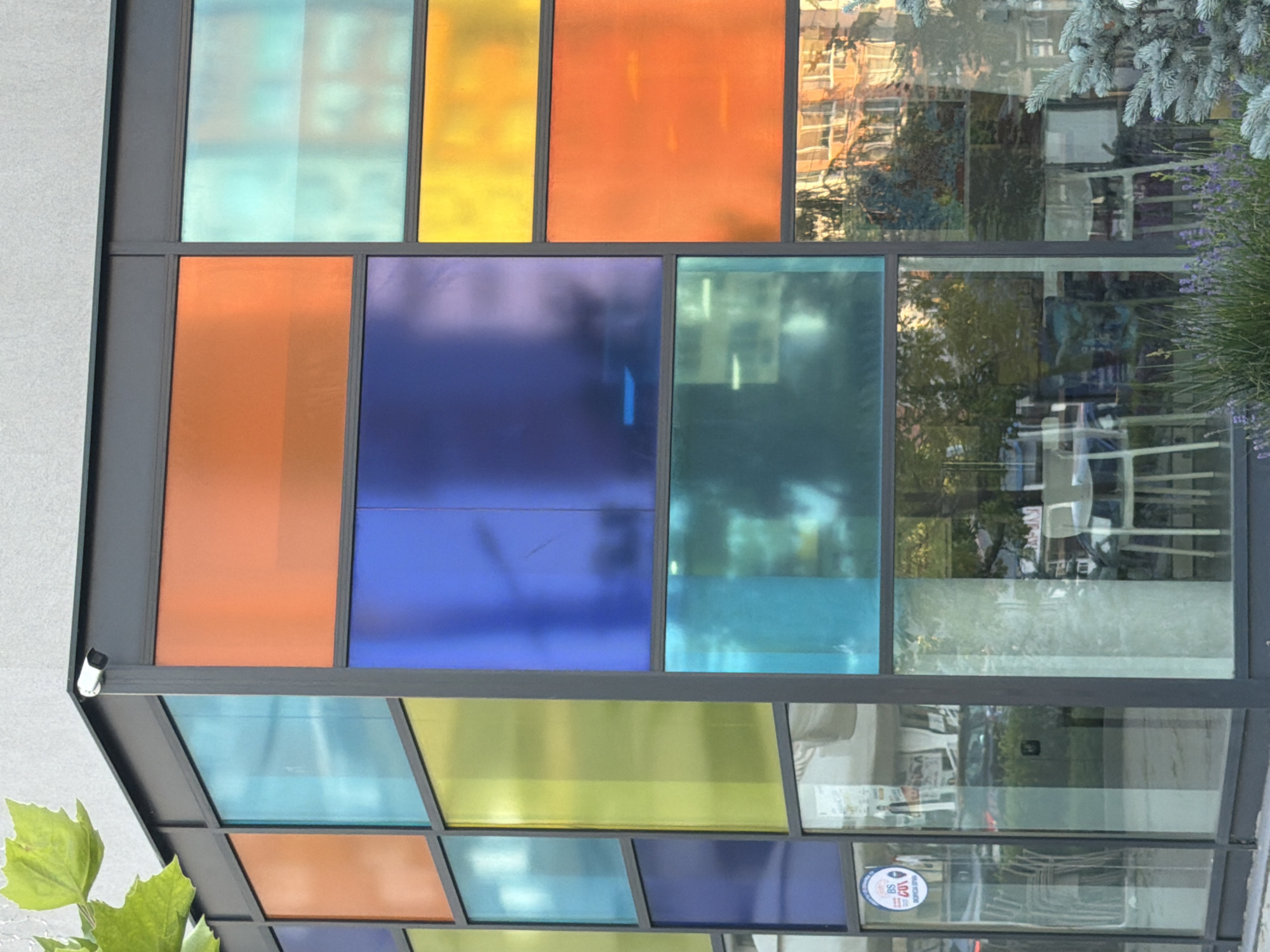 The image shows a modern building with a facade composed of large, colored glass panels in various shades of orange, blue, yellow, and turquoise. A security camera is visible mounted on the upper corner of the building. The glass panels are arranged in a grid-like pattern. Some of the panels are translucent, allowing for a blurred view of the interior and the reflection of the surrounding environment. Inside, tables and chairs are visible, suggesting a cafe or restaurant setting. The reflection on the glass shows trees and buildings. A small, circular sticker with text and an emblem is affixed to one of the lower glass panels on the left side. The scene appears to be in an urban environment, as indicated by the building structure and interior elements. The image is taken from a medium distance, showcasing the architectural features of the building.