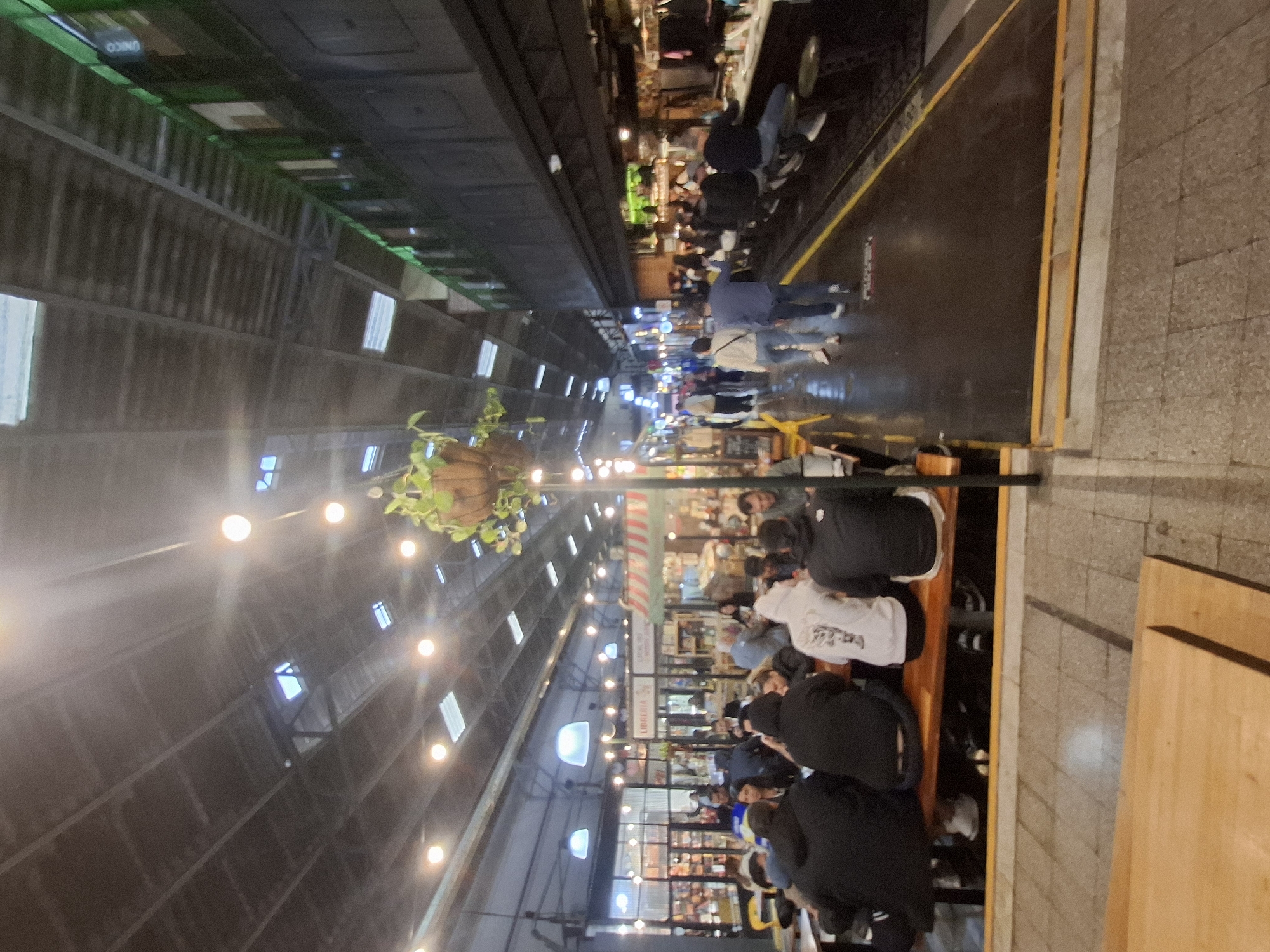 This image shows the interior of the San Telmo Market in Buenos Aires, Argentina. The view is lengthwise down the market aisle. A high, dark industrial roof with evenly spaced lights spans the length of the market. People are shopping, browsing, and eating in the market. There are vendors and customers along both sides of the aisle. The floor is shiny, suggesting it is wet or has been cleaned recently. Tables and chairs are visible to the right of the frame, suggesting a dining area.