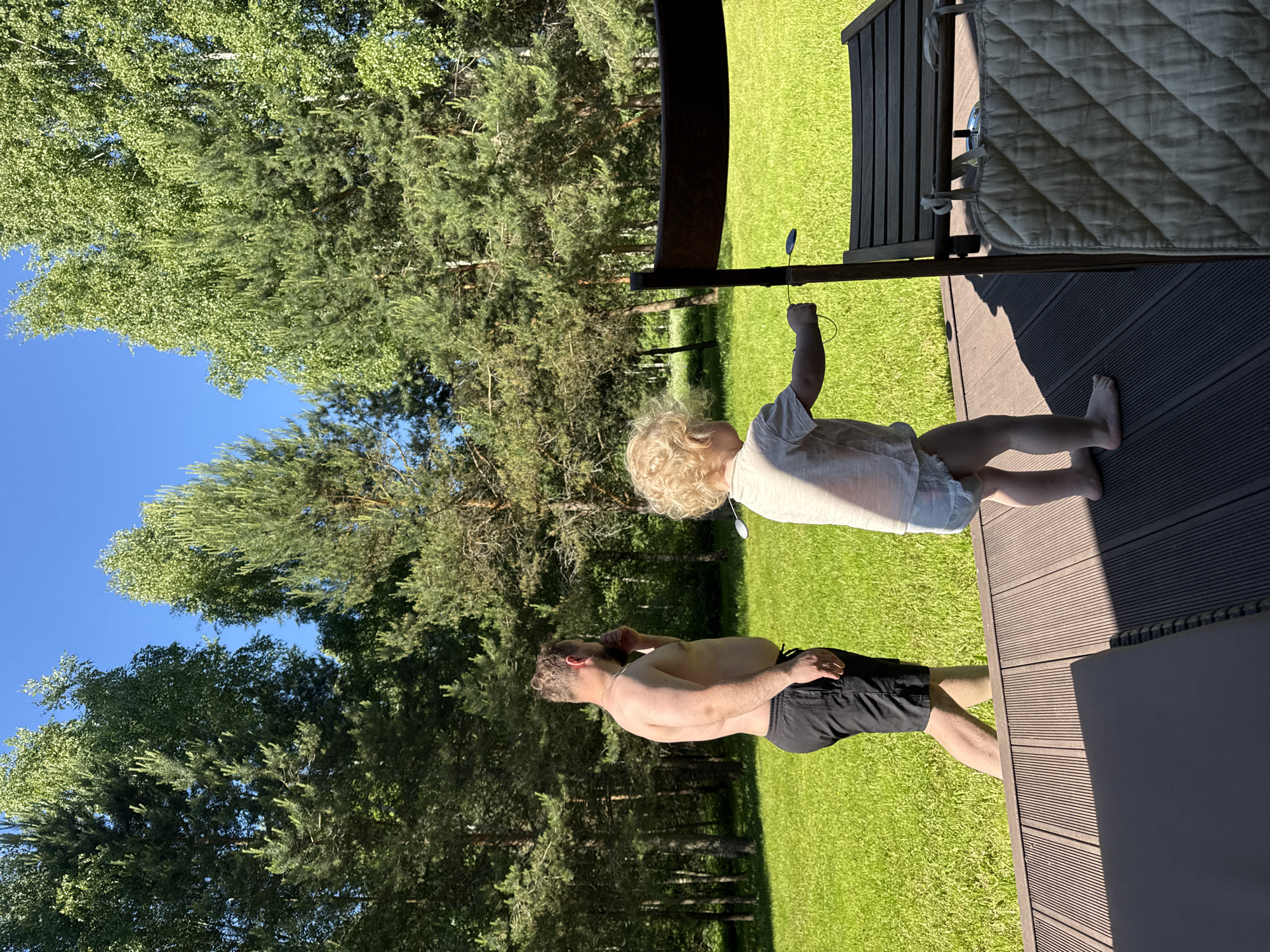 This image depicts an outdoor scene in Копнино, Russia, on a clear, sunny day. In the foreground, a dark slatted wooden deck extends across the bottom of the frame, with a partial view of a dark rectangular cushion or mat on the lower left. A small child, barefoot with curly blonde hair, stands on the deck in the right-center, facing away from the viewer. The child wears a light-colored t-shirt and a diaper, and holds a small, dark, spoon-like object attached to a string, extending it towards a dark wooden outdoor chair or table that partially obscures the child's lower body. A quilted, light-colored fabric item is hanging from the lower part of this wooden furniture.
Beyond the deck, a vibrant green lawn spans the midground. An adult male, shirtless with a beard and short dark hair, walks across the lawn from left to right, facing partially away from the viewer. He wears black athletic shorts and is holding a dark smartphone to his right ear, as if engaged in a call. A wedding ring is visible on his left hand.
The background consists of a dense treeline, featuring a mix of tall coniferous trees (pines or firs) and deciduous trees with lighter green foliage (birches). A clear, bright blue sky is visible above the treeline. The strong sunlight casts distinct shadows on the wooden deck.