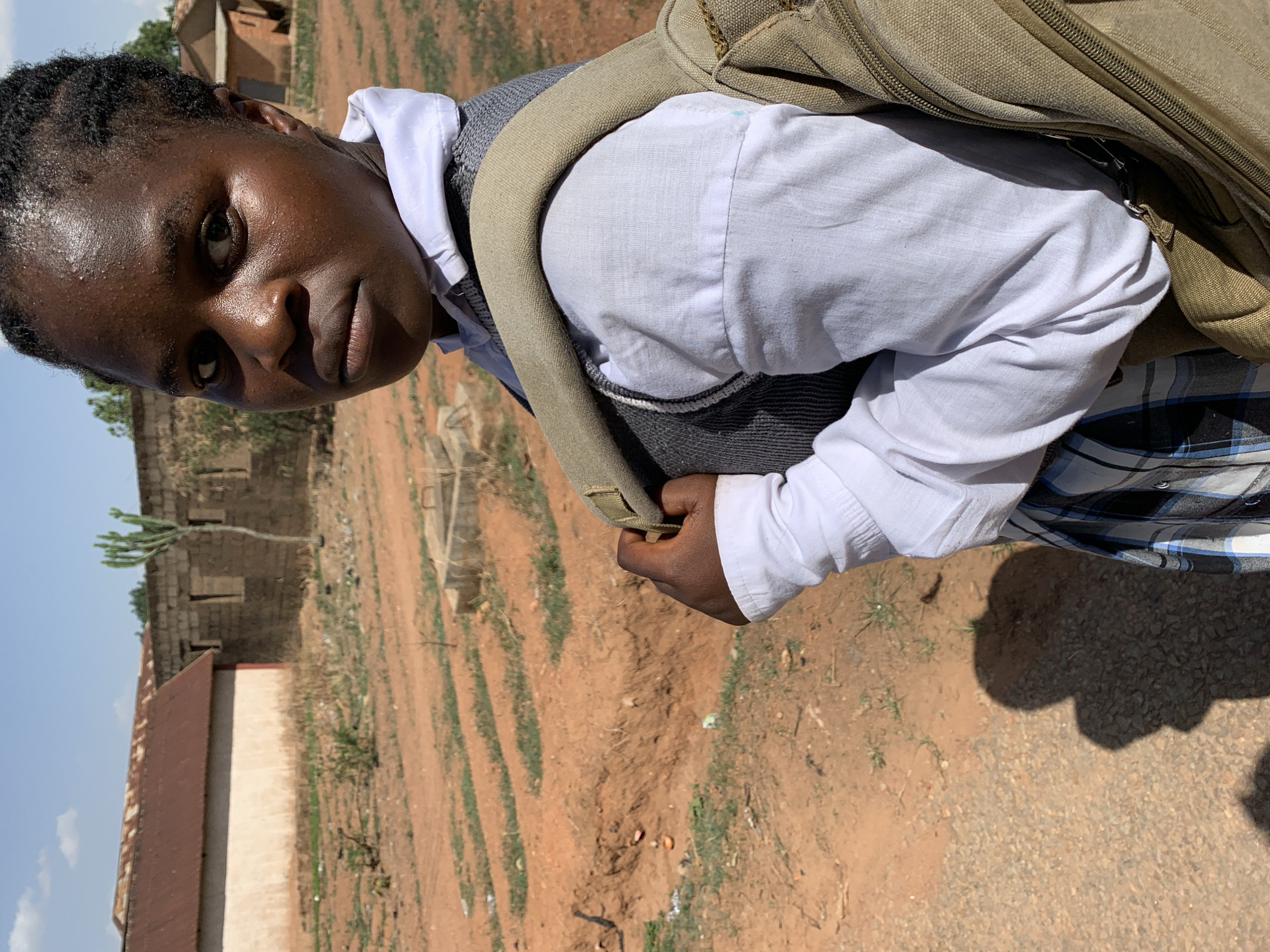 An adolescent girl with dark skin, dressed in a white shirt, grey vest, and plaid skirt, is carrying a khaki backpack in an outdoor setting in Vom, Nigeria. The ground is predominantly reddish-brown dirt with sparse green vegetation. A low building with a reddish roof is visible on the left side of the frame, while another building with unplastered walls and open windows is visible in the background. The sky is a light blue. The scene appears to be photographed on a sunny day.