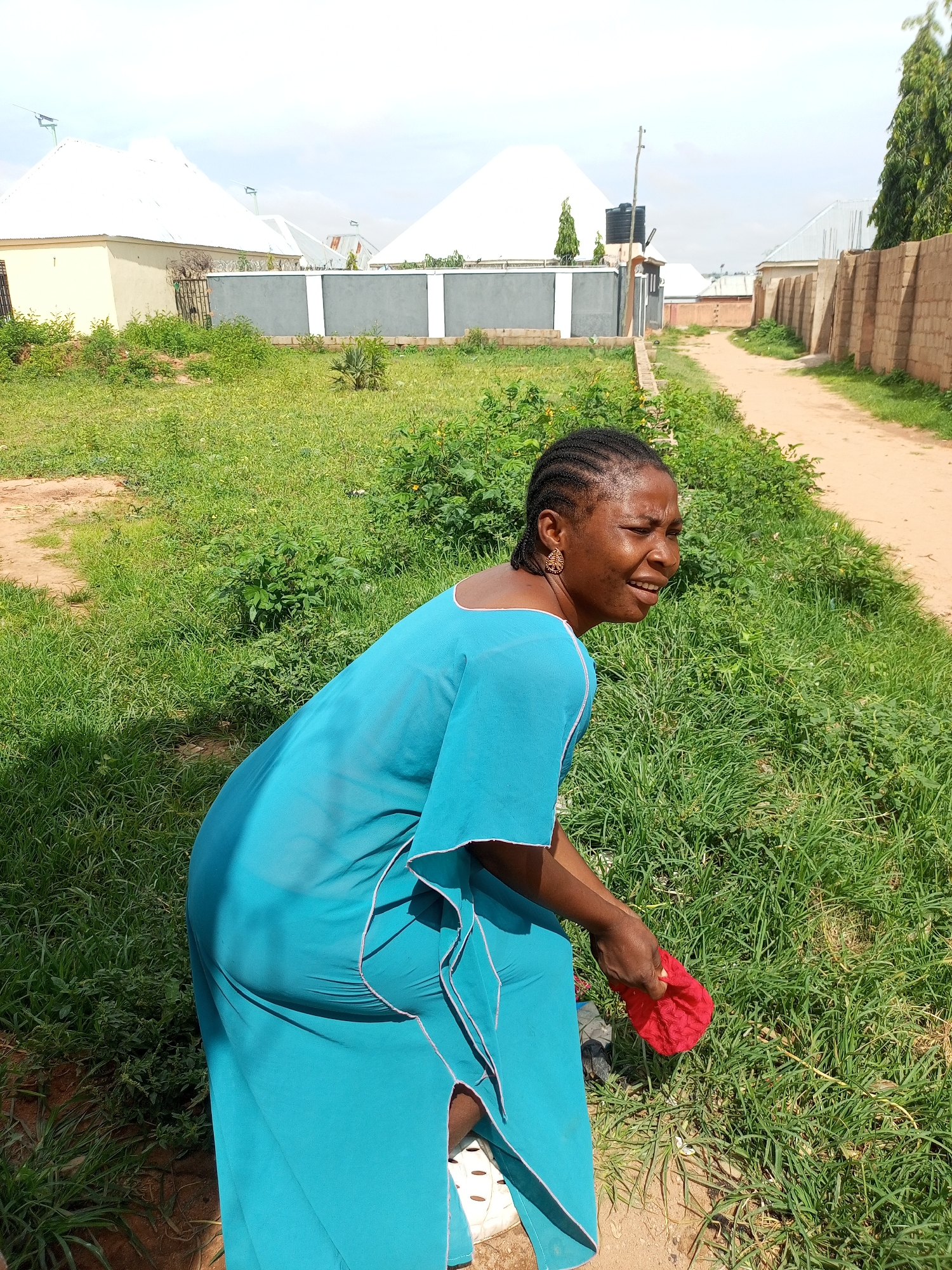 The photograph captures a full-body outdoor shot of a woman wearing a blue dress and white shoes. She is positioned to the left of the frame, facing away from the camera, with her head turned to the right, looking upwards and to the side with a somewhat distressed expression. Her hair is styled in braids, and she wears gold earrings. In her right hand, she holds a crumpled red object.
The woman is standing in a grassy area, with a dirt path visible to her right, leading into the distance. The background features several buildings with white roofs, some of which appear to be houses. A concrete wall with grey and white accents runs along the side of the property. Trees and vegetation are scattered throughout the scene. The overall environment suggests a residential area. The image is taken in the city of Tempure, Nigeria, likely in a suburban or semi-rural setting, given the mix of housing and open, undeveloped land.