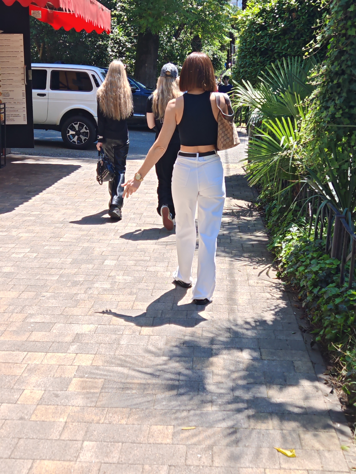 Three individuals are depicted from behind, walking away from the camera on a paved sidewalk on a bright, sunny day. The person closest to the viewer is on the right, wearing a black crop top and white, loose-fitting pants, accessorized with a gold watch on their left wrist and a beige and brown monogrammed shoulder bag.
To their left, the second person has long, wavy blonde hair and is dressed in black clothing, including leather-look pants, and carries a black studded bag in their left hand. Further in the background, a third person, also with blonde hair, wears a baseball cap and dark attire.
The sidewalk, paved with light-colored rectangular blocks, shows strong shadows from the people and overhead trees, indicating clear, sunny weather. On the left side of the path, a building with a red awning and an unreadable menu board is visible. A white SUV, resembling a Lada Niva, with distinctive black and silver rims, is parked near the building, with a dark car parked behind it.
The right side of the pathway is bordered by dense green foliage, featuring various bushes and large, palm-like plants, all contained behind a black metal fence. The scene suggests an outdoor commercial or recreational area, possibly a street or park path. No specific text is clearly legible in the image.