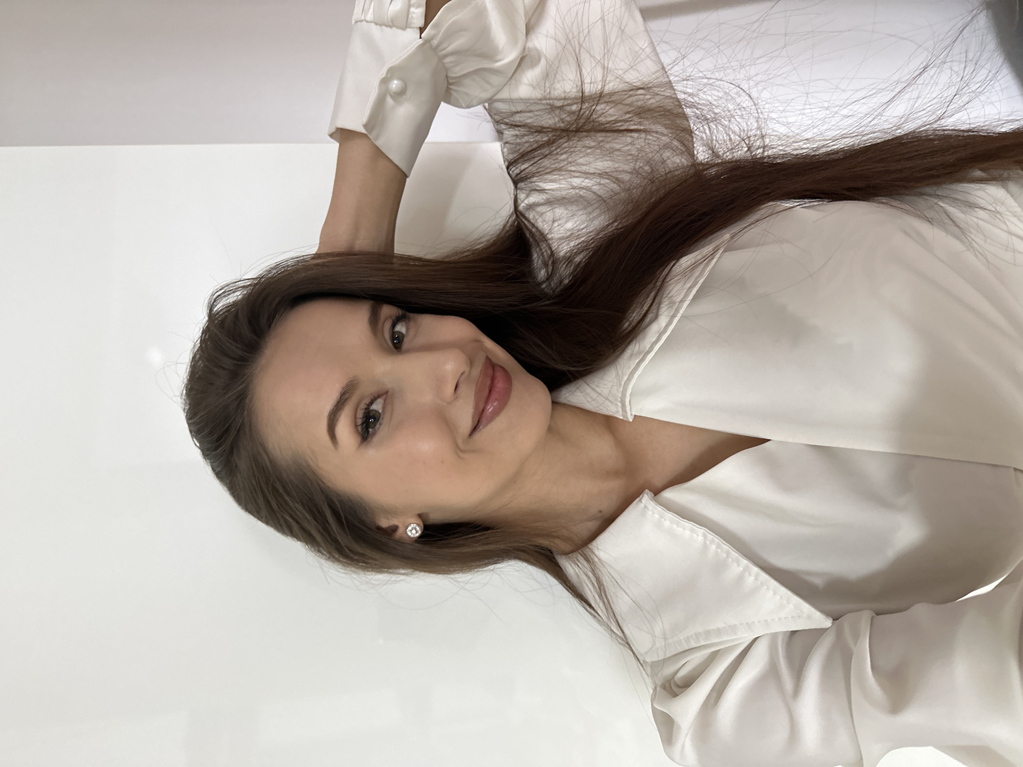 A woman is pictured in a close-up shot, rotated to the side. She has long, dark brown hair, light skin, and is wearing a white satin or silk blouse. Her arm is raised behind her head. She is wearing an earring. The background is white. There is no visible indication of the location being Yekaterinburg, Russia.