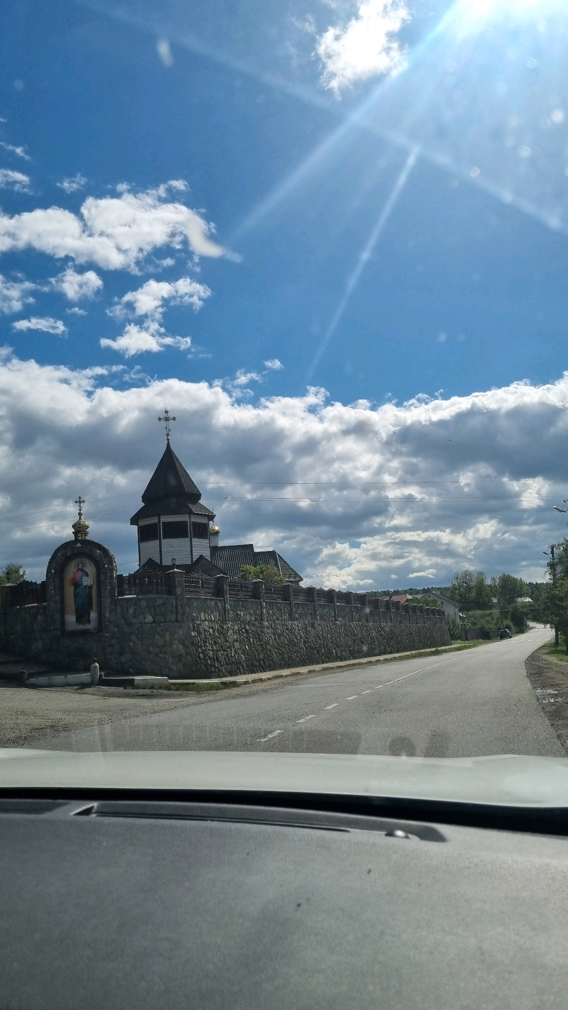 A wide-angle, eye-level, outdoor shot captures a view from inside a vehicle, looking out towards a rural road and a religious structure. The sky is a vibrant blue with scattered white clouds, and the sun is bright, casting strong light rays. In the mid-ground, a stone wall encloses a church with a dark, octagonal steeple topped with a cross. To the left of the church, a small arched monument features a mosaic image of a figure holding a book, also topped with a cross. The road curves to the right, leading into the distance past trees and houses. The foreground shows the interior of the car, with the dashboard and windshield visible. The scene is located in Bania Lysovytska, Ukraine.