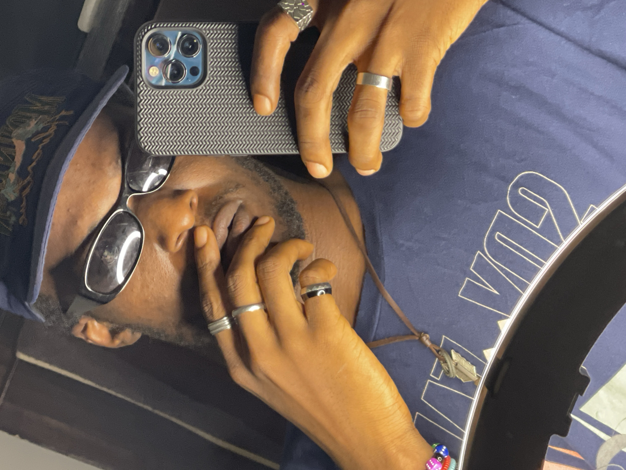 A young Black man wearing a dark blue baseball cap, oval-shaped sunglasses, and a dark blue t-shirt is taking a selfie. He has on several silver rings on his fingers and a necklace with a feather pendant. He is holding a dark grey textured phone in his right hand and has his left hand covering his mouth. The image appears to have been taken indoors with artificial lighting. There is no visible indication of the city of Abuja, Nigeria.