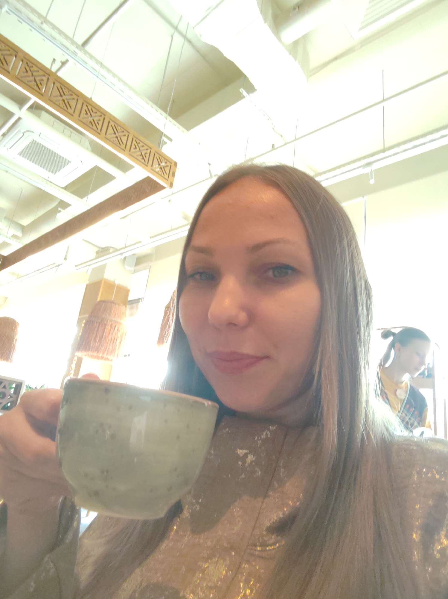 A woman with long blonde hair is taking a selfie while holding a ceramic cup in her left hand. She is wearing a shimmering gold top. In the background, another person with their hair in a bun is visible, wearing a yellow shirt and a patterned scarf. The environment appears to be indoors with bright lighting and exposed ceiling elements, possibly a cafe or restaurant. There are decorative hanging lamps with fringed shades in the background. The image does not provide any specific clues about the location being Syktyvkar, Russia.