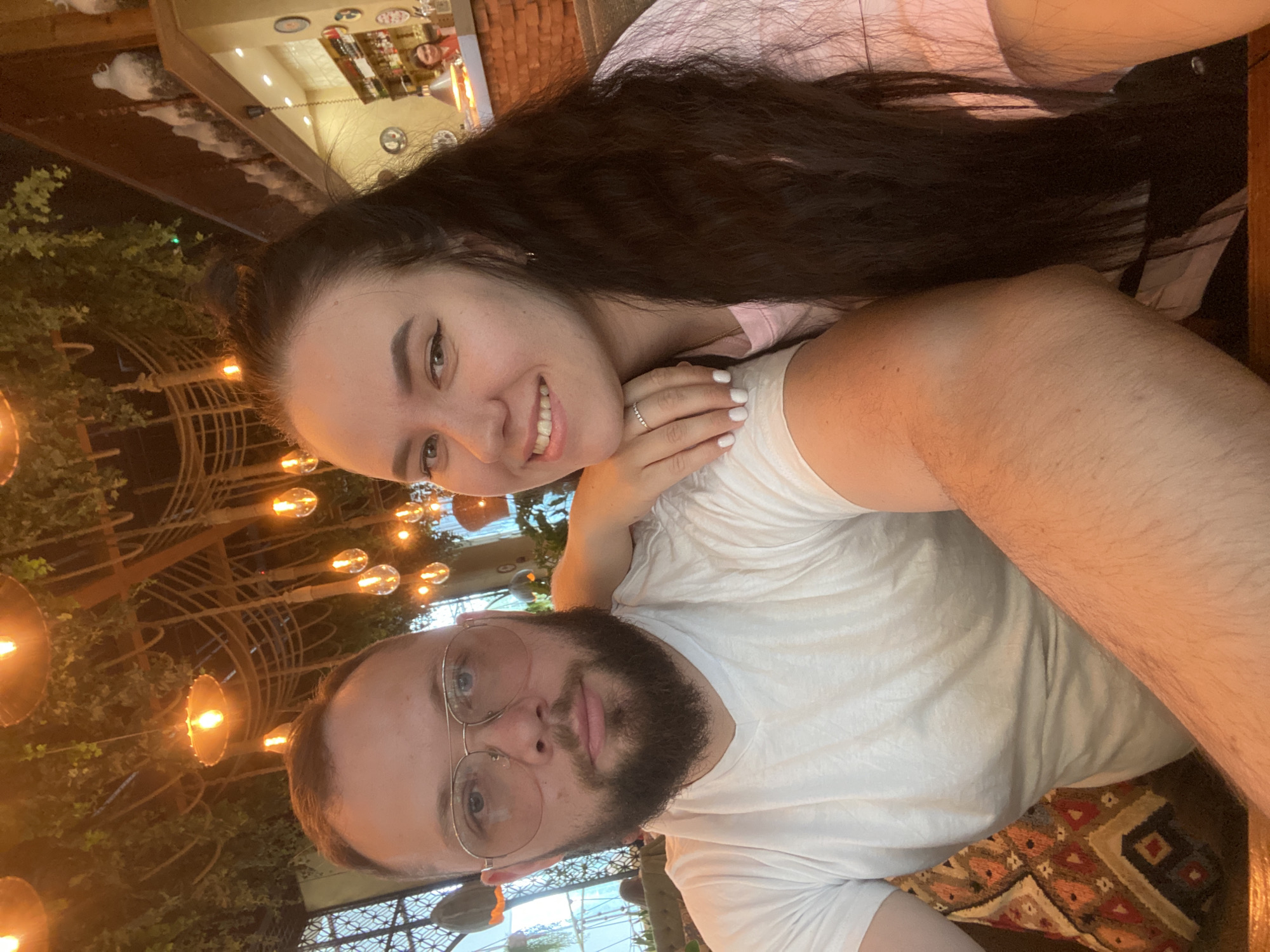 Two individuals, a woman and a man, are in close proximity, possibly in a selfie. The woman has long, dark hair and is smiling. She is wearing a ring on her finger. The man has a beard and is wearing glasses and a white shirt. They appear to be in a restaurant or cafe. The background features decorative lighting and greenery. The location is identified as Perm, Russia.