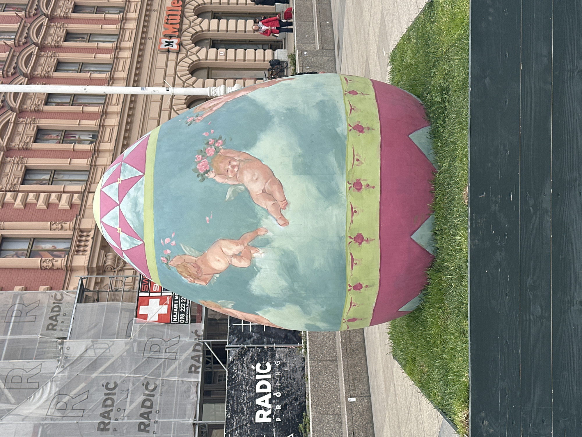 Here is a forensic description of the image:
The image shows a large, decorated Easter egg in an outdoor public space in the city of Zagreb, Croatia. The egg's surface is painted with a scene of cherubic figures in a classical style, against a light blue backdrop. The top and bottom portions of the egg are decorated with pink and green geometric patterns.
Behind the egg, scaffolding and banners with the text "RADIĆ PRONG" are visible, partially obscuring a building. The building has a sign that reads "Müller" indicating a business establishment. People are seen in the background near the building.
The egg is positioned on a patch of grass, next to a grey walkway and a dark-stained wood structure. The setting appears to be a city square or plaza.