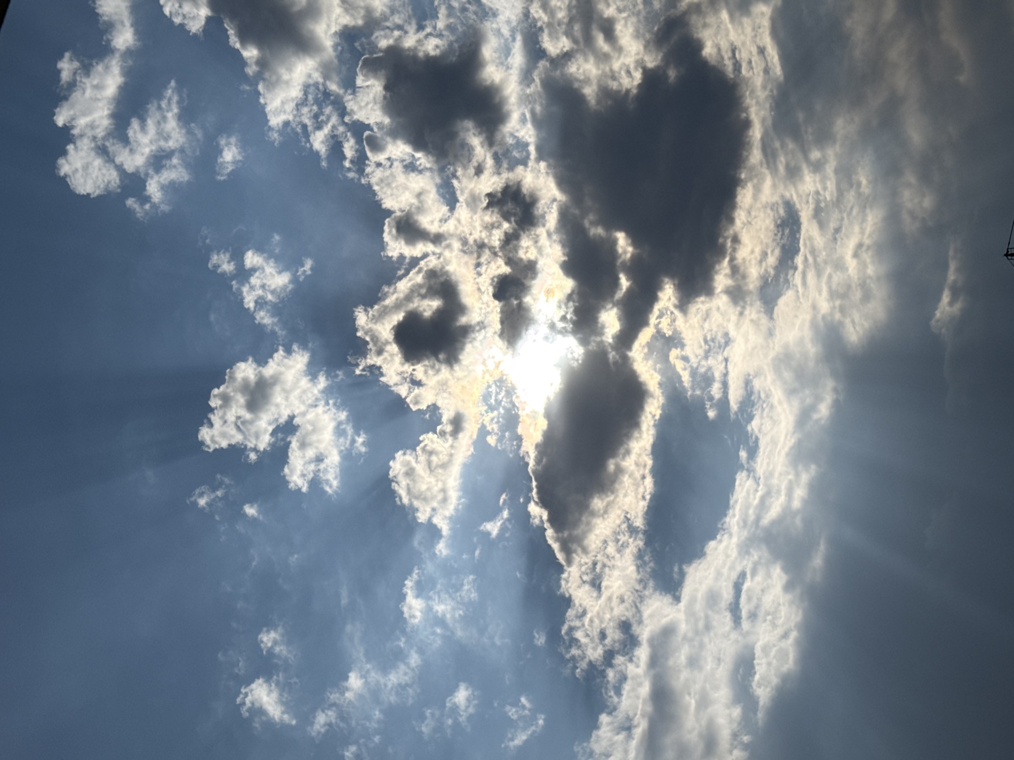 An outdoor, vertical, medium shot captures a dramatic sky with sun rays breaking through dark clouds. The sun is partially obscured by thick, dark gray clouds in the center of the frame, with bright white and light gray cumulus clouds scattered throughout the blue sky. Sunbeams emanate from behind the central clouds, casting streaks of light across the scene. The lower portion of the image is dominated by darker, denser clouds, with subtle rays of light piercing through them. A faint silhouette of a crane or tower-like structure is visible at the very bottom edge of the frame. The scene appears to be a natural phenomenon of a cloudy sky during daylight.