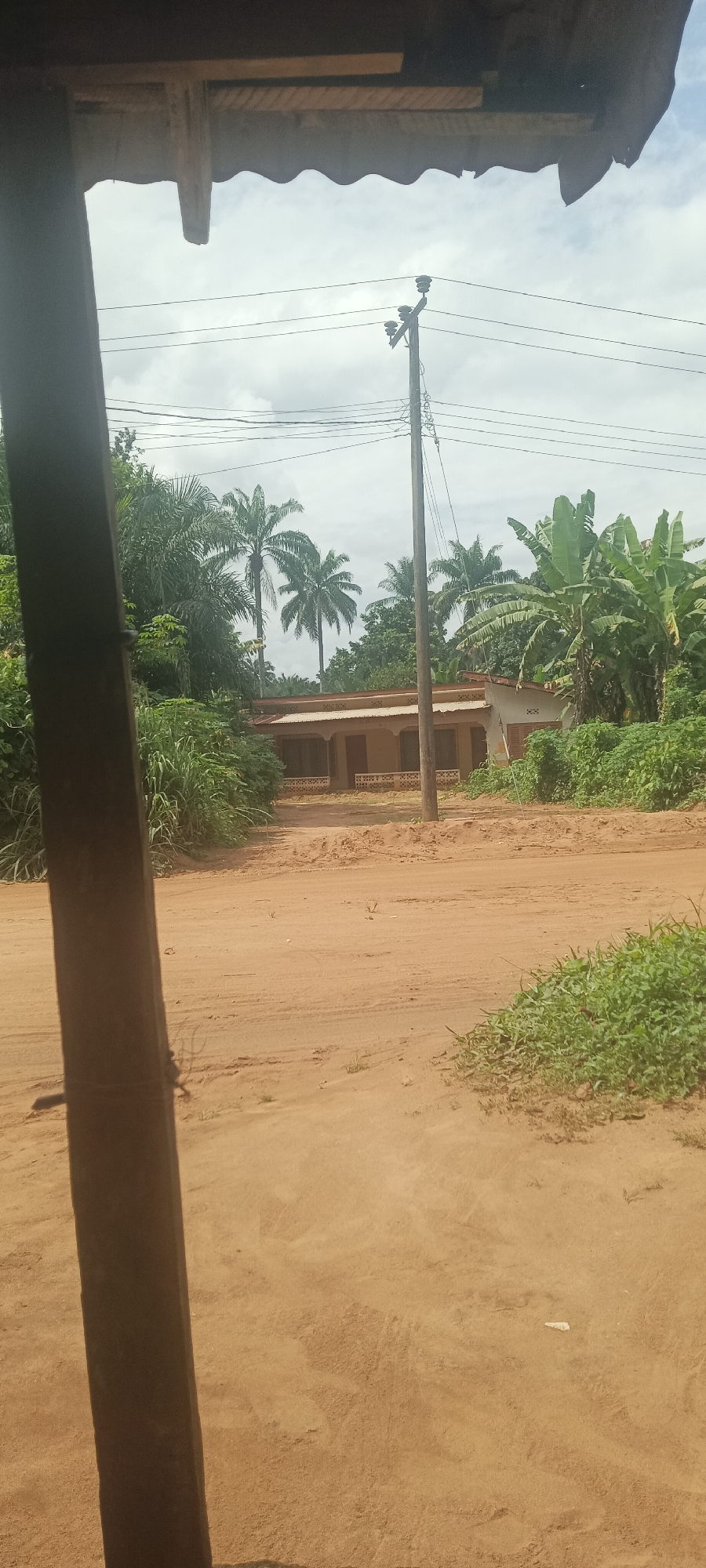 The image shows a dusty, unpaved road in Amauzu, Nigeria. A small, single-story building with a veranda sits at the end of the road, surrounded by lush vegetation including palm trees and banana plants. A wooden utility pole with power lines is visible in front of the building. The photograph is taken from under the overhang of a rudimentary structure with a wooden support beam partially visible in the foreground. The sky is overcast. There are no people visible in the image.