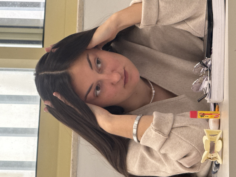 A young adult female is depicted in a close-up, chest-up shot, seated indoors at a light-colored table. She has long, dark brown hair with a center part and is wearing a light beige V-neck sweater, a pearl necklace, and a silver-toned bracelet with rectangular links on her left wrist. Her hands are positioned with fingers touching her temples, and her gaze is directed downwards and to the right. On the table directly in front of her, from left to right, are a light yellow hair claw clip, a yellow tube of ChapStick with a red cap and strawberry branding, a pair of dark-framed eyeglasses resting on a stack of white papers or books, and a partially visible dark-colored smartphone. The background shows a blurred interior, including a light-colored wall and a window with vertical elements and a yellow frame. The specific location within Tel-Aviv, Israel, is not discernible from the visual evidence.