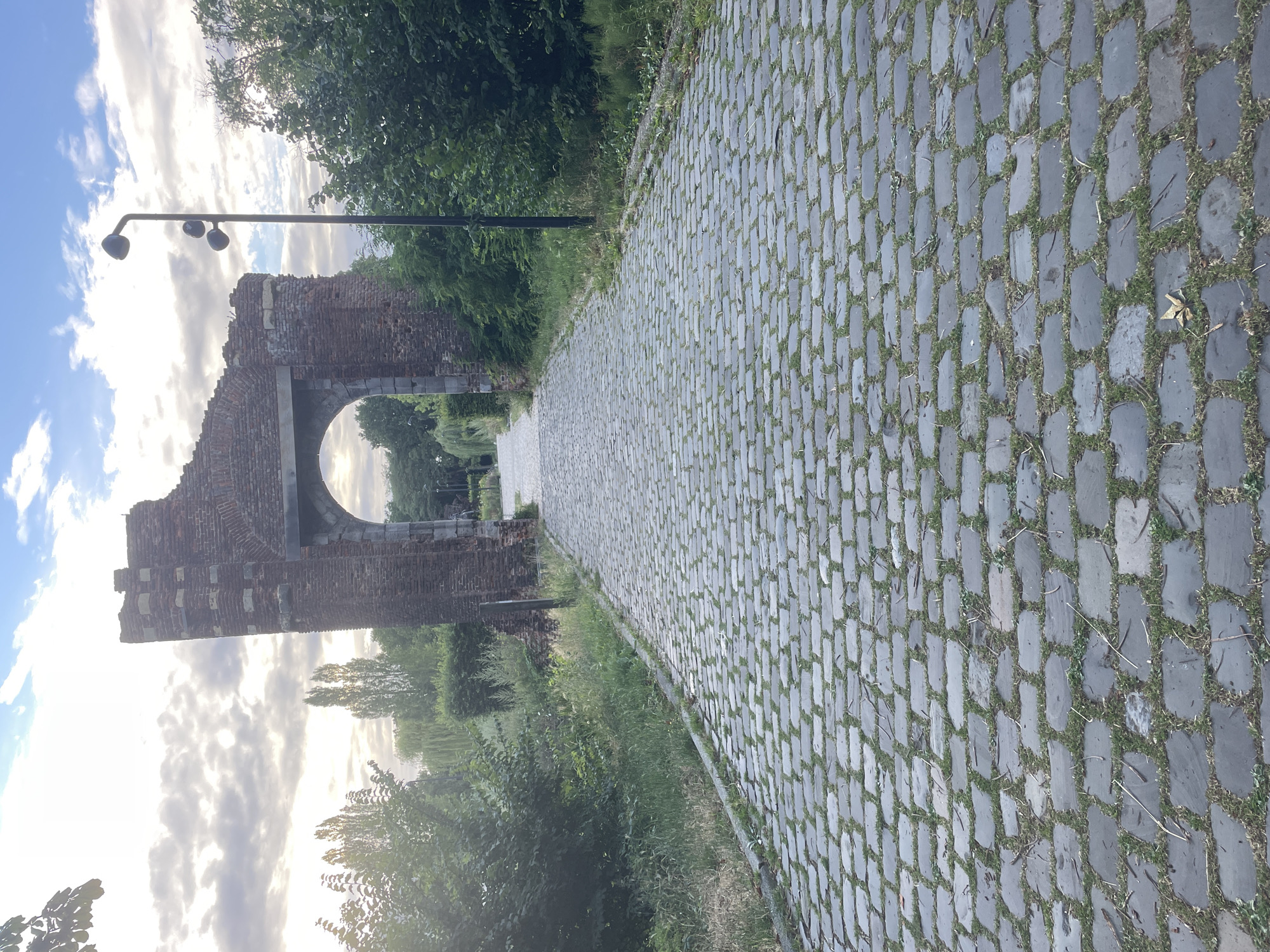 The image captures a stone-paved pathway curving through a green park-like setting in Weert, Netherlands. The pathway, laid with grey, rectangular stones interspersed with grass, dominates the foreground and recedes into the distance. To the left of the path, a crumbling brick archway stands as a historical remnant, possibly part of an old structure, with some supporting columns visible. The archway is weathered, showing signs of age with missing bricks and exposed stone. Beyond the arch, trees and foliage form a lush backdrop, with hints of more greenery and a clear sky with scattered clouds suggesting it's either morning or late afternoon. A street lamp with three lights is positioned near the archway, indicating a public space. The overall mood is serene and slightly melancholic, with the historical structure evoking a sense of the past within a natural, tranquil environment. No people are visible, and there is no visible text.