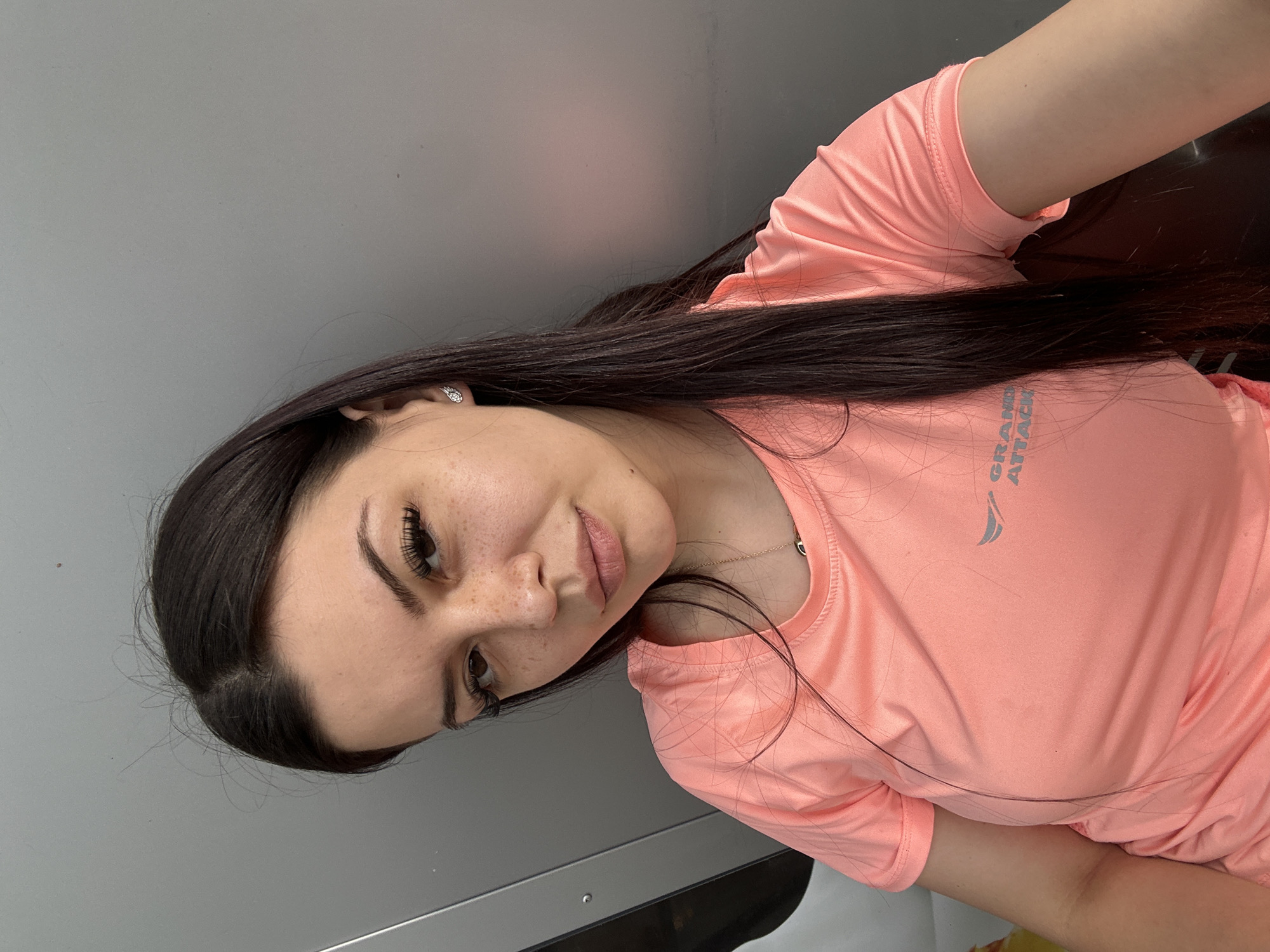 A young woman with dark hair and fair skin, wearing a coral-colored t-shirt, is pictured in an indoor setting. Her hair is long and straight, with some strands falling across her face and shoulders. She has prominent eyelashes and a natural makeup look, highlighted by freckles across her nose and cheeks. She is wearing a delicate necklace with a dark pendant and small, sparkly earrings. The background appears to be a plain, metallic gray wall, suggesting an urban or industrial environment. The lighting is soft, casting subtle shadows on her face and clothing. The t-shirt has a gray logo on the left chest area, partially obscured, which appears to be text and a graphic element. The image is a close-up portrait, focusing on the woman's upper body and face. The location of Sliven, Bulgaria is not determinable from the visual information provided in the image.