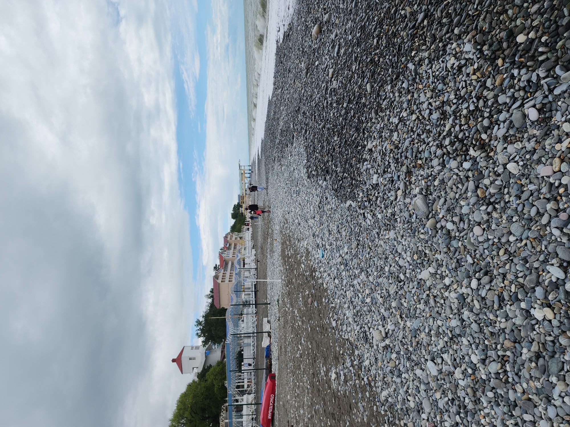 The image shows a beach in Sochi, Russia. The beach is composed of small, grey stones. The sea is on the right side of the frame with white foam of the waves, and the sky above is partly cloudy. On the left, there are buildings, trees, beach umbrellas, and lounge chairs. A few people are seen walking in the middle ground of the image. A red life-saving boat with writing in Cyrillic is visible near the bottom left of the image.