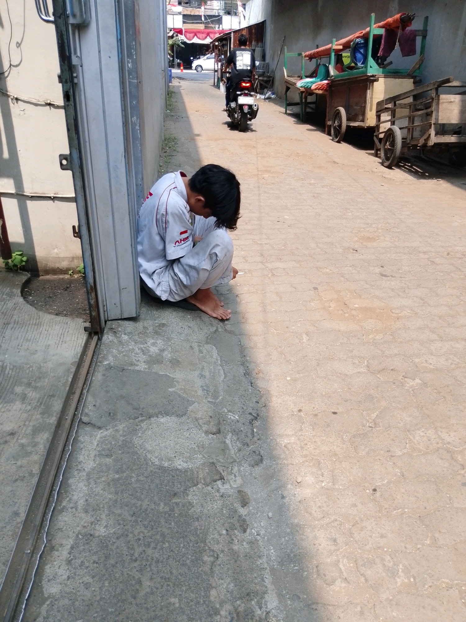 The image captures a scene on a sunlit street in Pasuruan, Indonesia. In the foreground, a young person in a light-colored uniform with red trim is seated on the ground, facing away from the camera and hunched over. Their head is bowed, and their feet are visible. The person is positioned next to a tall, gray metal structure that appears to be part of a building or a gate.
Behind the seated individual, the street extends into the distance. A person on a black motorcycle with a visible license plate is riding away from the viewer. To the right of the street, there are several wooden carts and stalls with various items, including what appears to be fabric or tarps in shades of red and green. The background shows buildings with red awnings. The ground is paved with concrete and appears somewhat weathered, with patches of discoloration and debris. The overall environment suggests a commercial or residential street with activity.