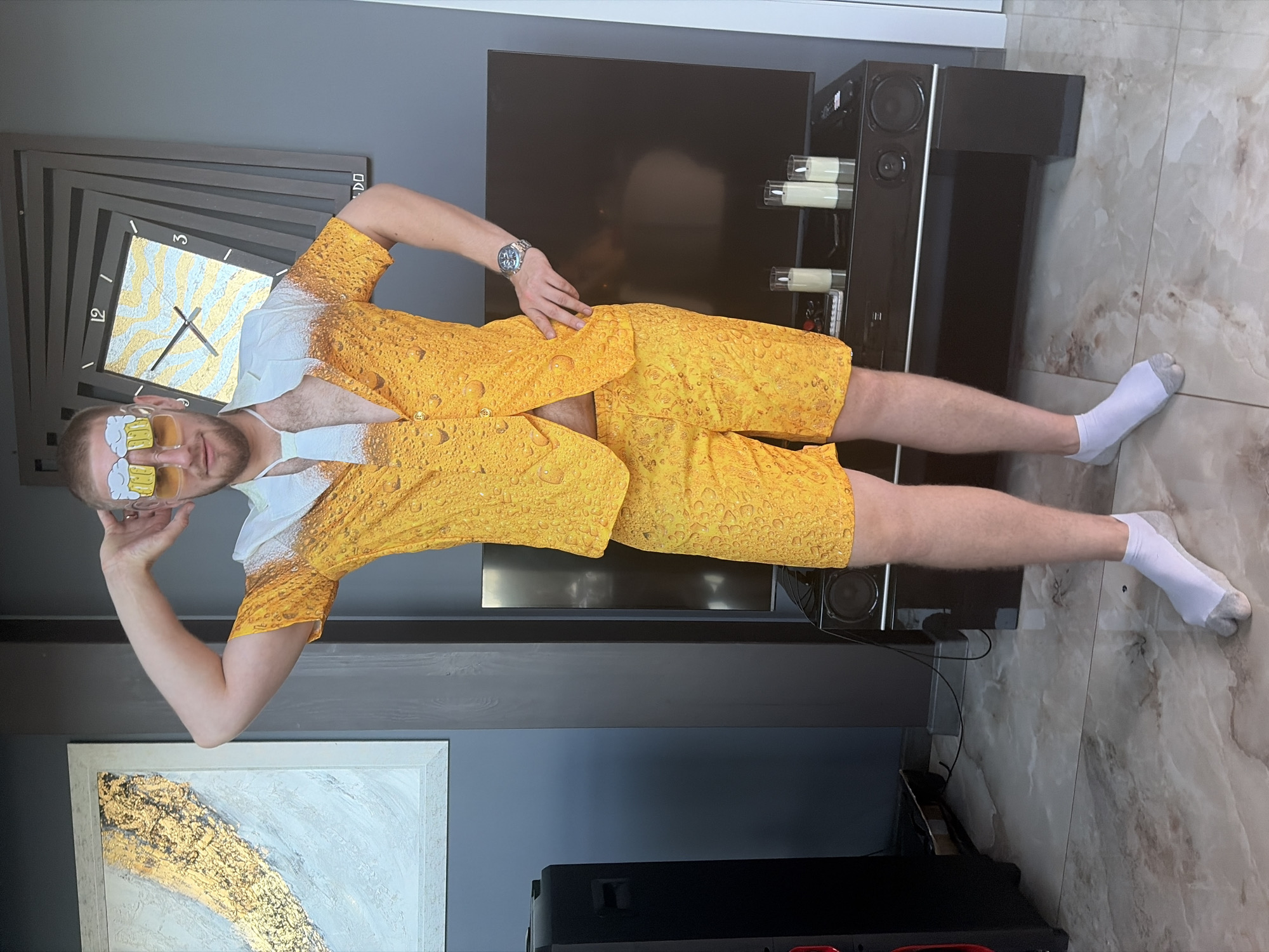 A man stands in what appears to be a living room or entertainment area, wearing a novelty outfit designed to resemble beer with frothy tops. He has his left arm raised behind his head and is looking towards the camera with a slight smile. He is wearing yellow sunglasses with beer mug shaped decorations. His outfit consists of a short-sleeved button-down shirt and matching shorts, both patterned with yellow and white bubbles, simulating beer. He has a white tie draped around his neck and is wearing white split-toe socks. In the background, a large flat-screen television is visible, with a sound system and decorative candles on a stand below it. To the left of the television are large black speakers. A framed piece of art with gold and white abstract patterns hangs on the wall to the man's left. To his right, a modern clock with a spiraling frame is mounted on the wall. The floor is tiled with a marble-like pattern. The overall impression is of a staged photo, likely for social media or a humorous occasion. The man is Caucasian, with short hair and a light beard. He is wearing a watch on his left wrist. The lighting suggests an indoor setting, possibly during the daytime. The room appears to be modern and well-decorated.