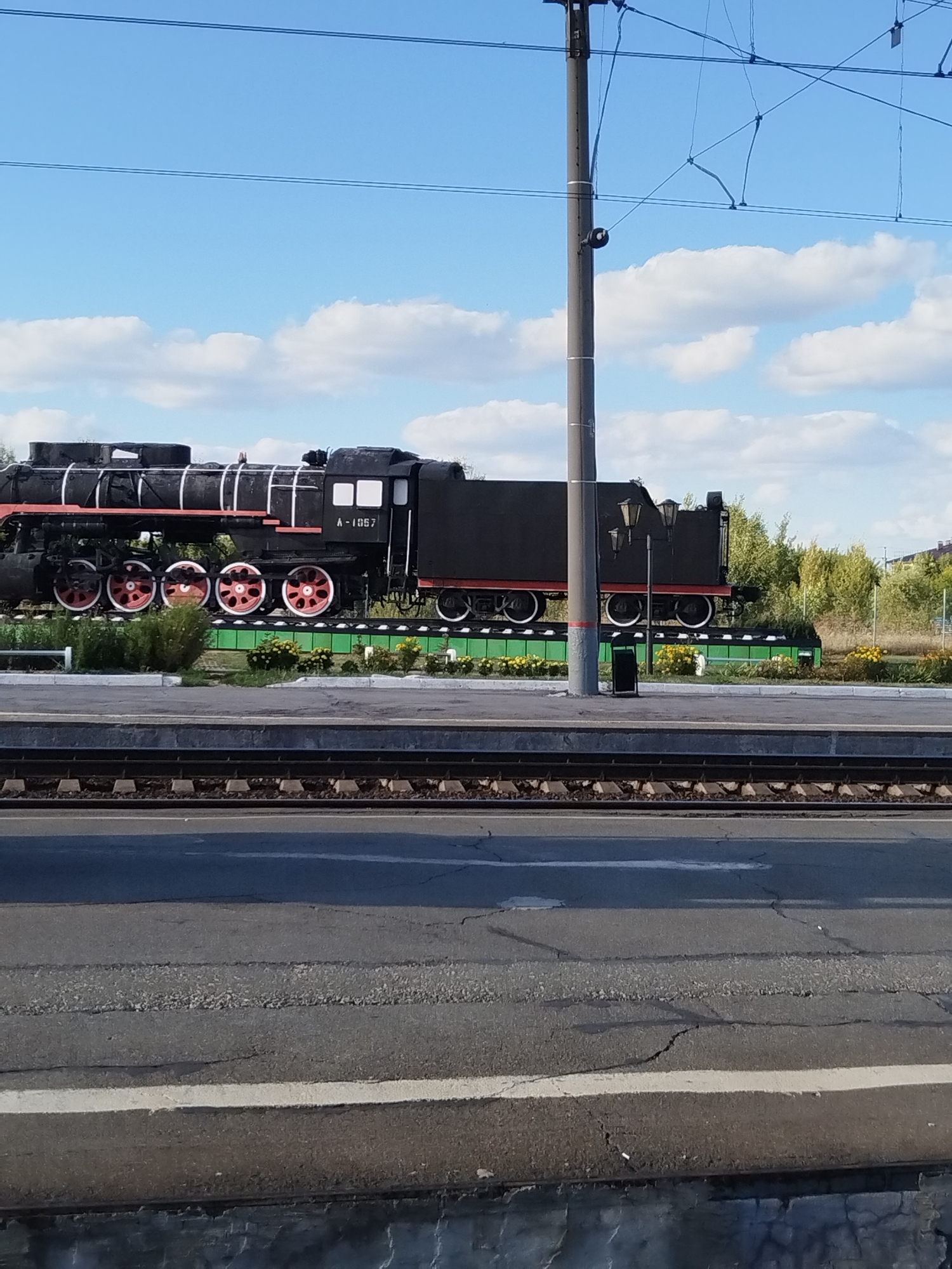 The image shows a black steam locomotive on display, identified by the number "A-1067" painted on its side. The locomotive is positioned on a display track, bordered by a green platform and an assortment of yellow and green flowers. Behind the locomotive, trees and buildings are visible under a partly cloudy blue sky. In the foreground, there are train tracks and a paved area. The scene is likely a public space in Barabinsk, Russia, possibly a park or a station exhibit, given the historical nature of the locomotive and the presence of train tracks. There are no people visible in the image.