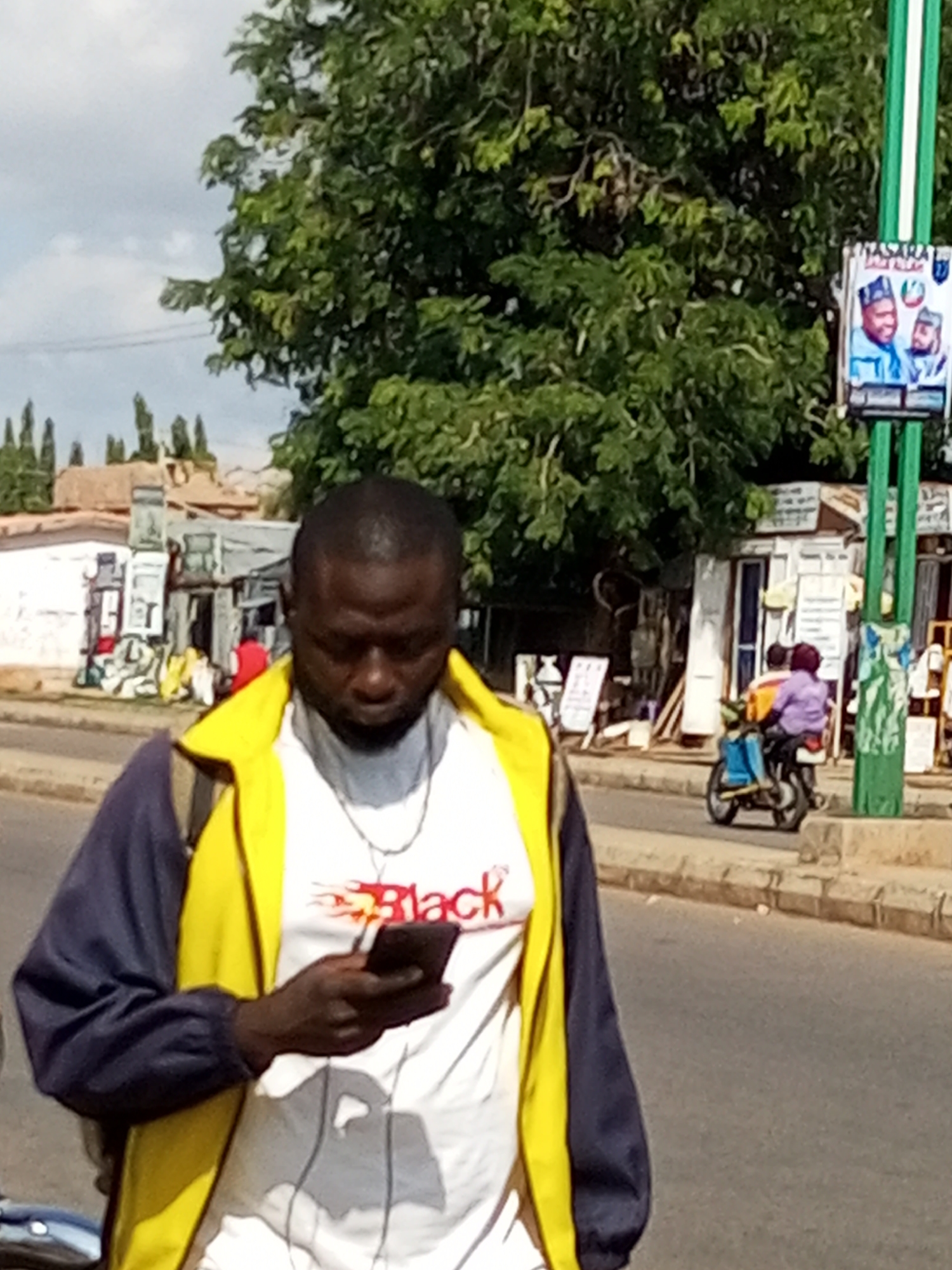 A medium shot shows a Black man in the foreground, looking down at a smartphone he holds in his right hand. He is wearing a white t-shirt with red lettering that reads "Black" with a flame graphic and a blue jacket with a yellow collar. He is walking on a paved street, with a sidewalk to his right. Behind him, there are several small shops and buildings, and a large tree with green leaves. A poster of two men in traditional clothing is visible on a green pole to the right of the frame, and a person on a motorcycle is seen in the background. The scene is set in the city of Gombe, Nigeria, possibly near a commercial area given the presence of shops and street advertising. The overall lighting suggests it is daytime, with some shadows present.