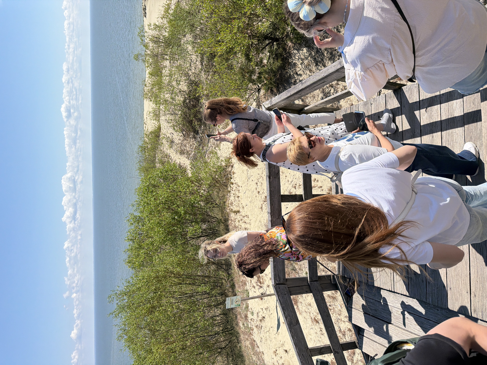 A group of people stand on a wooden boardwalk overlooking a sandy dune landscape and the sea under a blue sky with scattered clouds. A sign with an image of a person walking is visible on a pole to the left of the boardwalk. Several individuals are holding smartphones, suggesting they are taking pictures or videos. The people are dressed in casual summer clothing, and the overall atmosphere appears to be one of tourism or leisure. The location is Morskoye, Russia, likely a coastal area with sand dunes.