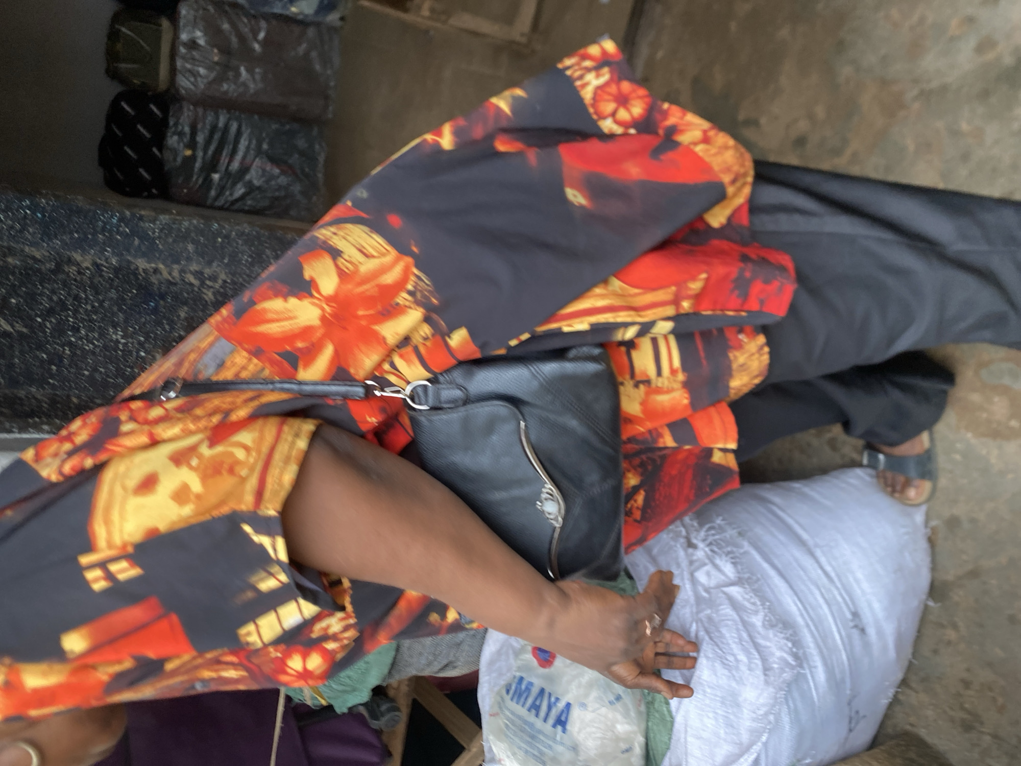 In Bukuru, Nigeria, a person stands next to a large white sack. They are wearing a black, red, and yellow patterned top and black pants. A black purse is slung over their shoulder. The environment appears to be an outdoor market or storage area, with stacks of goods visible in the background. The sack next to the person has some blue writing on it. The person's left hand is resting on the sack.