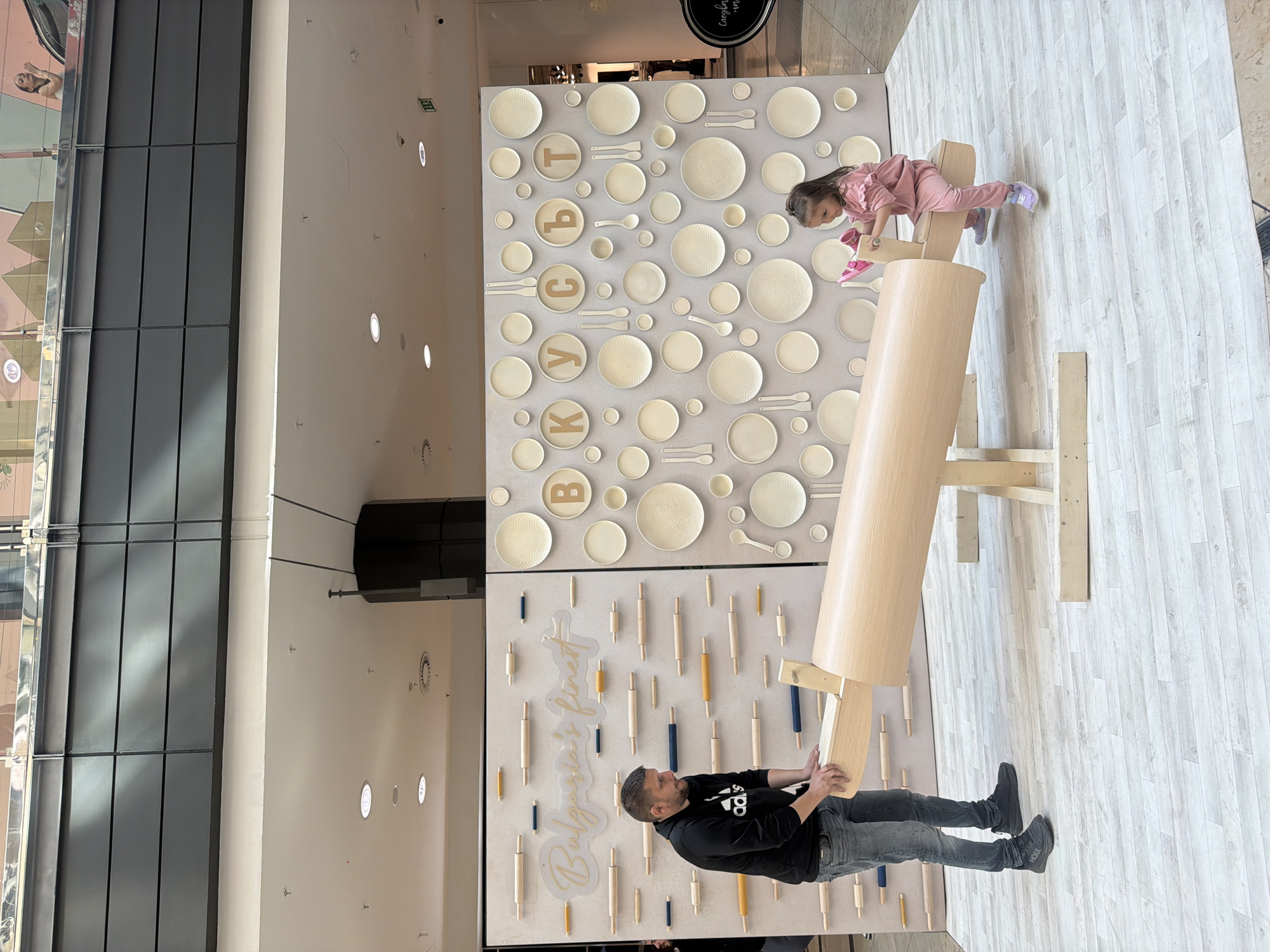 The image captures an interactive art installation inside what appears to be a shopping mall in Sofia, Bulgaria. A man is seen manipulating a large, wooden rolling pin-like structure that is part of the installation. A young girl is positioned atop the rolling pin, engaging with a section of the art piece featuring plates and utensils arranged on a wall. The installation's backdrop showcases two distinct panels, one adorned with plates and Cyrillic letters and another with rolling pins. The interior architecture features glass panels and a high ceiling, suggesting a modern, indoor environment.