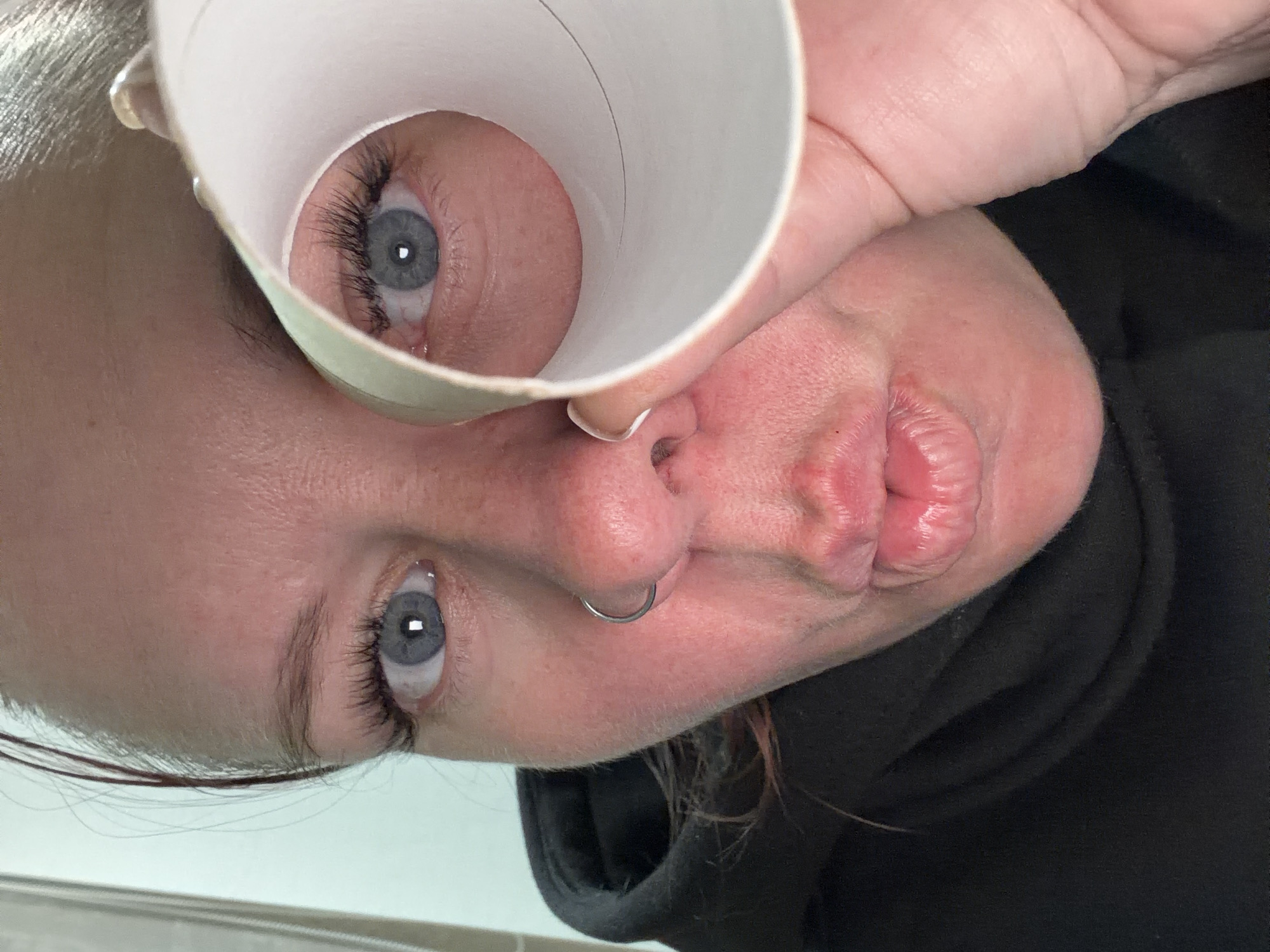 A close-up, tilted selfie depicts a person holding a white cardboard tube up to their right eye. The eye, viewed through the tube's opening, is gray-blue with dark eyelashes. The person's face is flushed, with a nose ring and pursed lips. They are wearing a black hooded sweatshirt. There's no information to determine if the subject and location are in the city of Chaikovsky, Russia.