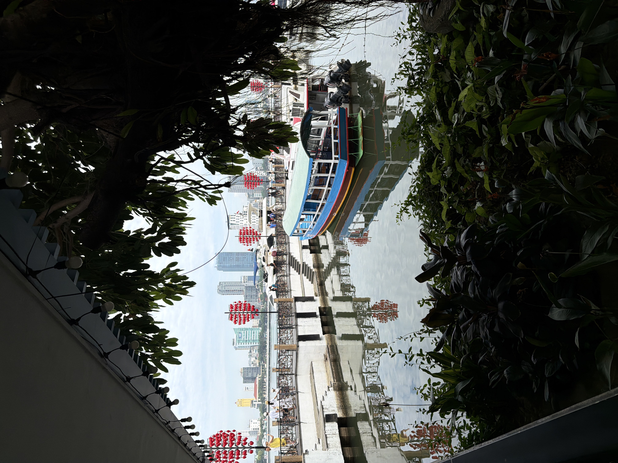 This image depicts a waterfront scene in the city of Đà Nẵng, Vietnam, likely near the Han River. In the foreground, lush green foliage partially obscures the view. A large tree branch with hanging roots extends from the right side, casting shadows.
The middle ground features a walkway alongside the water. Several people are visible on the walkway, some standing and others walking with bicycles. Decorative red heart-shaped lanterns are mounted on poles along the railing. A white structure with tiered steps leads down to the water.
Dominating the water are two boats. The closest boat is blue and green with a light-colored roof and appears to be a passenger boat. It is equipped with two outboard motors at the stern. Behind it, another larger boat, predominantly white with red accents, is moored. The buildings of the Đà Nẵng cityscape form the backdrop, with several modern high-rise buildings and other structures visible. The sky is overcast. The water reflects the surrounding structures, boats, and lanterns. The scene suggests a public area with recreational and sightseeing activities.