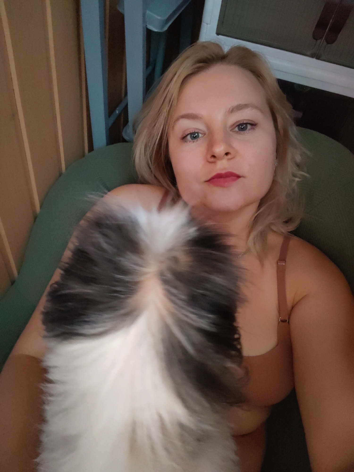 A Caucasian woman with blonde hair and blue eyes is shown in a close-up selfie. She is wearing a light brown bra and is holding a furry animal, possibly a cat or dog, in front of her face. The animal has black, white, and gray fur. The woman appears to be indoors, with a green chair and wooden walls visible in the background. The lighting suggests it is daytime. No specific location in Izhevsk, Russia, can be determined from the image.