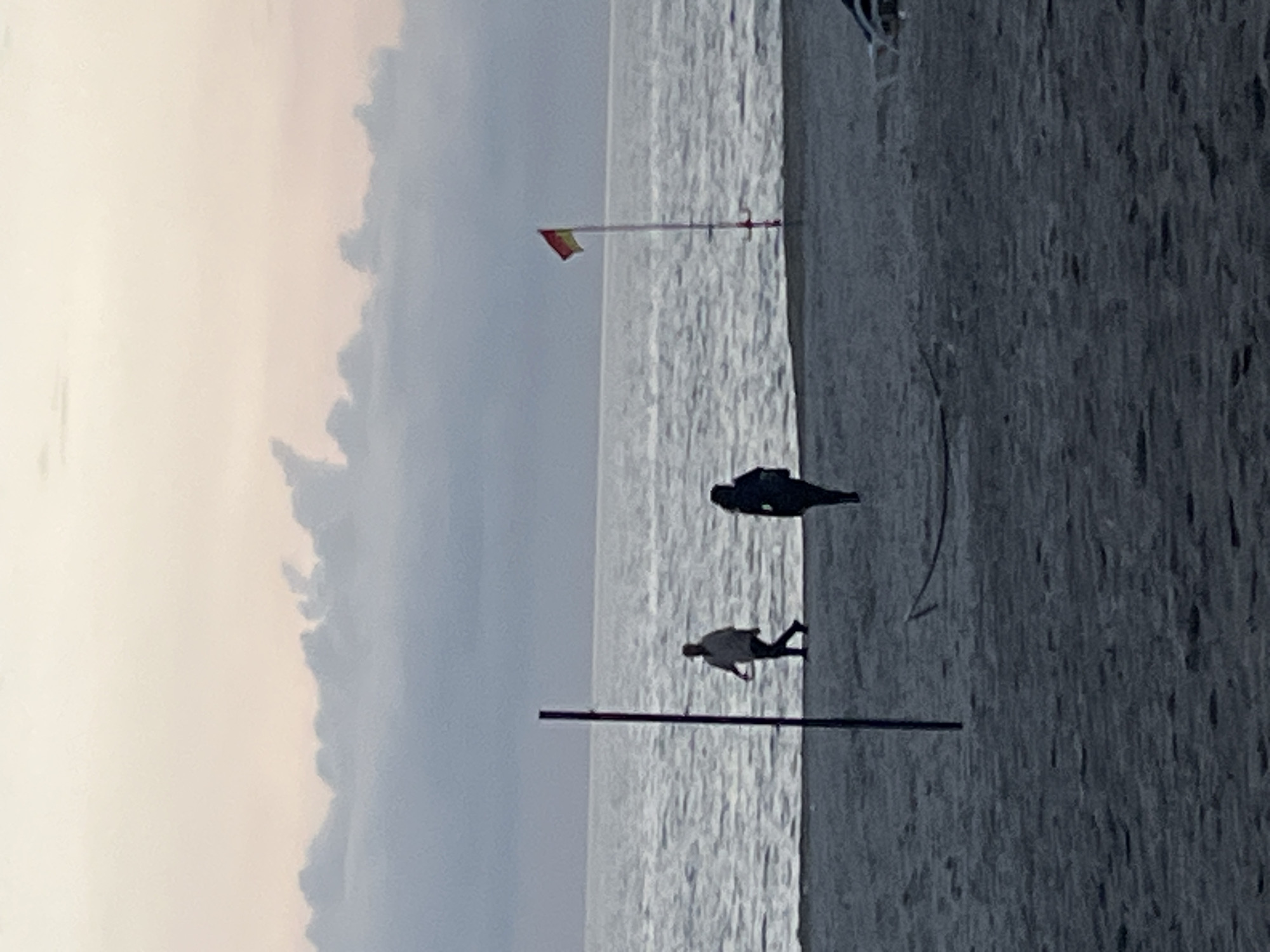 The image shows a beach scene. There is a cloudy sky to the left of the frame, and the sea extends to the center of the frame. A beach and possibly part of a structure are to the right. Two silhouetted figures are on the beach. A red and yellow flag is mounted on a pole. Another pole is visible closer to the bottom of the frame. The image appears to have been taken in Alanya, Turkey, judging by the flag.