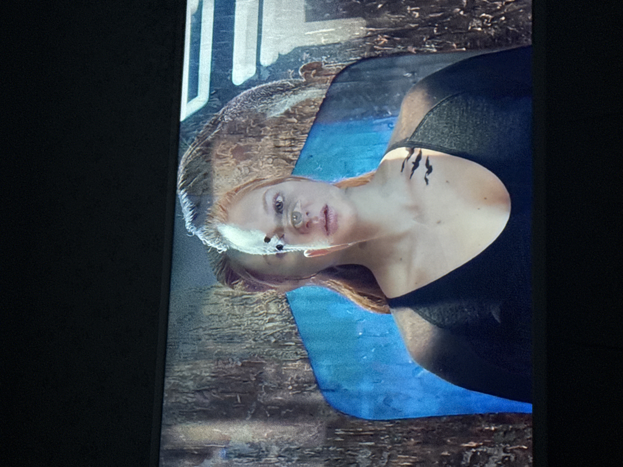 The image shows a woman with fair skin and reddish-blonde hair, wearing a dark top. She has what appears to be dark markings on her neck and chest. Her face is visible with a neutral expression. There is a blue-colored area behind her, with a wood-like texture beneath the blue. Part of a white letter is visible in the upper left corner, suggesting it is part of a poster. The location information provided is Mytishchi, Russia.
