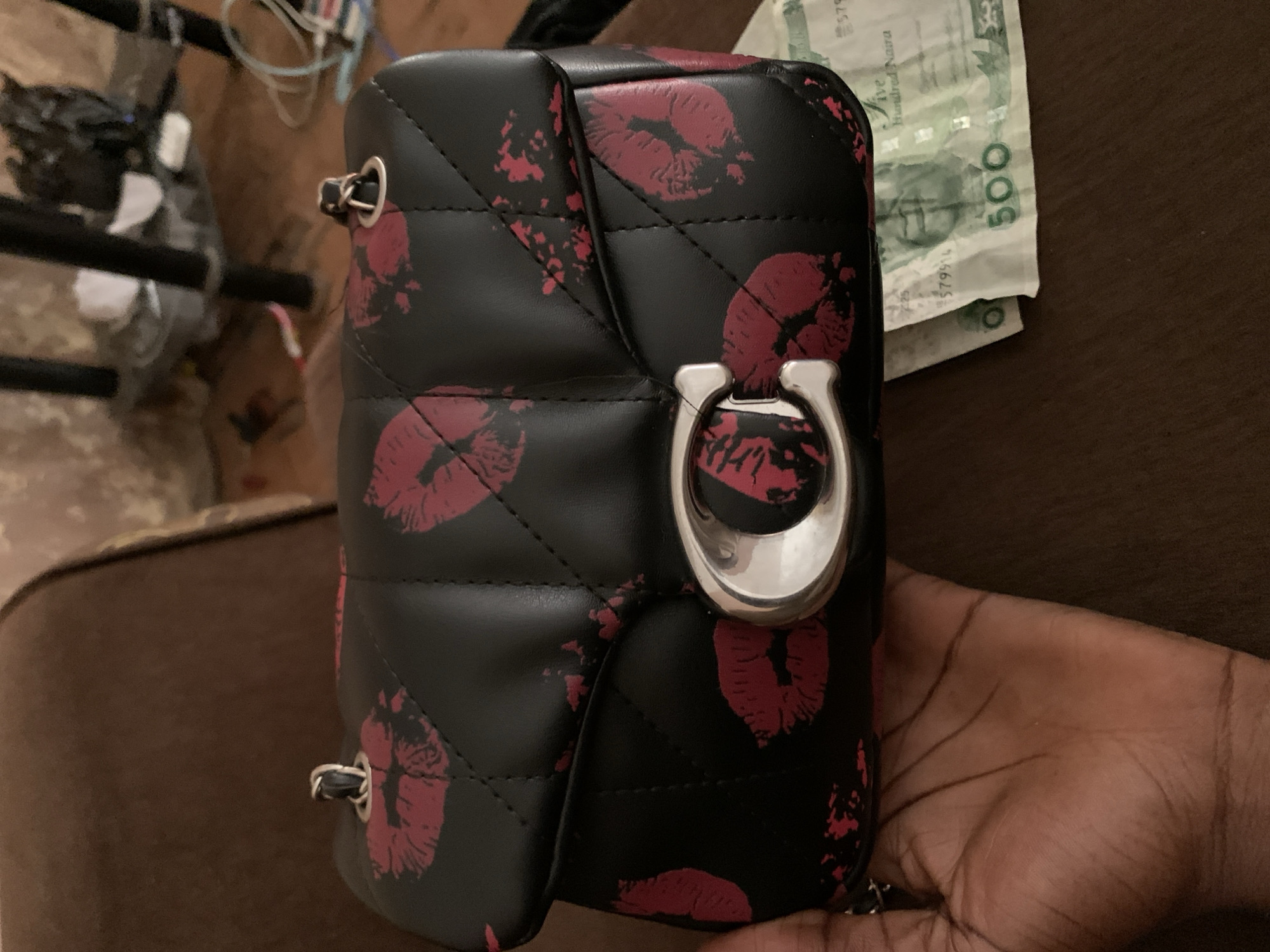 A black quilted handbag with a red lipstick kiss pattern is held by a dark-skinned person's hand. The handbag features a silver "C" logo on the front. Partially visible in the lower right corner are two Nigerian Naira banknotes, one showing "500". The background is a blurred brown surface, likely a couch or chair, with some out-of-focus objects in the distance suggesting an indoor environment in Jos, Nigeria.