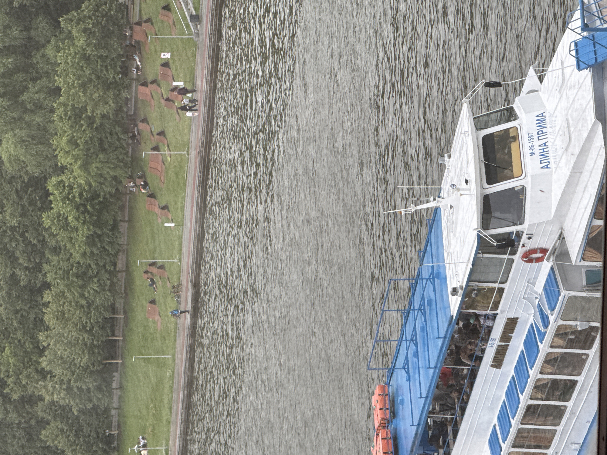 This is a photograph taken from an elevated perspective, looking down at a body of water and a waterfront area with trees and a boat. The scene appears to be taking place outdoors during the daytime, under what seems to be overcast skies given the muted lighting.
The waterfront is lined with lush green trees. In the foreground on the left, there are rows of brown benches or loungers on a grassy area, some of which are occupied by people. Further along the bank, several bicycles are parked.
A white boat with blue accents is docked on the right side of the image, floating on the water. The boat has multiple windows and a visible upper deck. It appears to be carrying passengers, with many people visible through the windows. There are also orange life preservers attached to the side of the boat.
The water is choppy, with visible ripples and waves. The overall atmosphere seems to be a leisurely day at the waterfront, possibly a park or a recreational area.
Visible text on the boat includes "М-06-1597" and "АЛИНА ПРИМА" written in blue. The location context provided is Moscow, Russia.