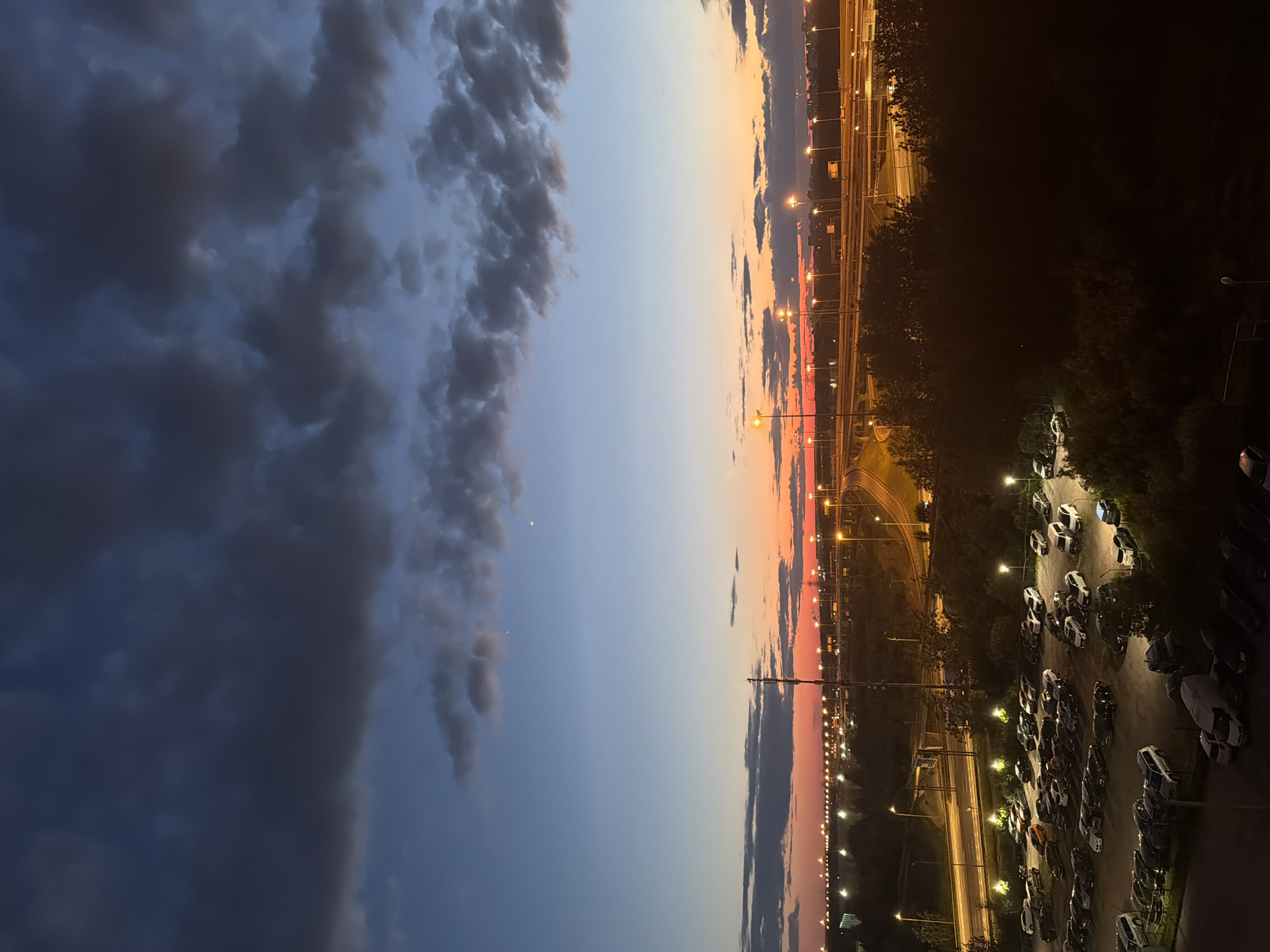 An aerial, high-angle, long shot captures a parking lot filled with cars, a highway overpass, and a dusky sky over a city. The sky exhibits a gradient of colors, from deep blue at the top to orange and red near the horizon, with scattered clouds. Streetlights illuminate the highway and portions of the parking lot. The parking lot contains numerous vehicles of various makes and models, parked in rows. Trees and foliage partially obscure some of the parked cars and the landscape. In the distance, the city skyline is visible, marked by a multitude of illuminated buildings and lights. The overall impression is one of an urban landscape at dusk.