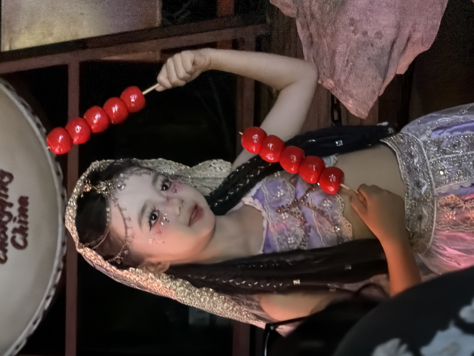 The image shows a young person in a traditional Chinese-inspired outfit, adorned with intricate beadwork and a delicate headdress. They are holding a skewer of candied hawthorn berries, a popular Chinese street food. The person has glitter makeup around their eyes, adding a touch of festivity. They are lying on their side, propped up on their arm, and looking towards the viewer with a playful expression. The background is dark and indistinct, suggesting an evening or indoor setting, possibly at a night market or festival. The overall mood is cheerful and whimsical. Visible text on a round object in the foreground reads "Choi Hung China." The setting appears to be Yuzhong District, China.