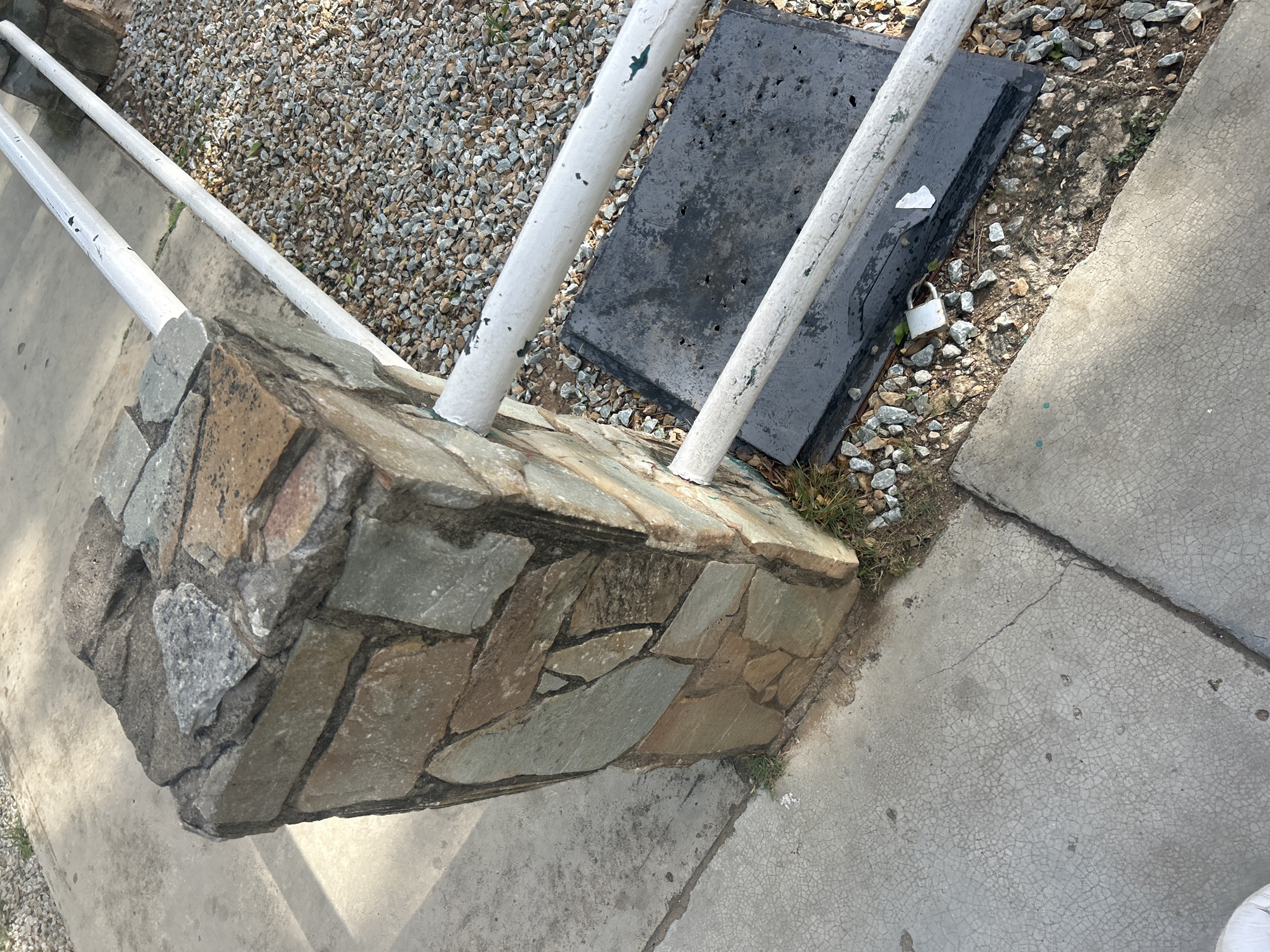 A stone pillar supports a white metal railing. The pillar is constructed from irregularly shaped stones in various earthy tones, including gray, brown, and tan, mortared together. The railing consists of two horizontal white pipes, one above the other, showing signs of chipping and wear. Beyond the railing, a ground surface of gravel is visible, interspersed with some dry grass. To the left of the pillar, a concrete surface with visible cracking and some small dark spots is present. The scene appears to be outdoors, with shadows suggesting it is daytime. There are no people visible in the image. The specific location in the city of Santa Ana, Venezuela, is not discernible from the visual information provided.