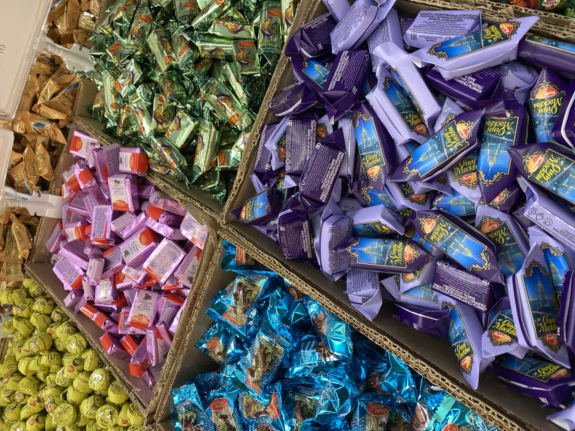 The image displays a close-up view of several cardboard boxes filled with an assortment of candies. The candies are individually wrapped in colorful foil and paper packaging.
In the top-left box, numerous candies are wrapped in shiny green foil, each featuring an illustration of a rooster. Adjacent to this, in a larger box, are candies with purple wrappers. These wrappers have a decorative design depicting a city skyline, possibly St. Petersburg, and the text "Огни Москвы" (Lights of Moscow) is prominently displayed. Further down, a box contains candies wrapped in light purple and pink foil. Some of these wrappers show images of grapes and text in Russian, such as "ЗОЛОТОЕ СУФЛЕ" (Golden Soufflé). To the right of the light purple candies, another box is filled with candies in bright blue wrappers, also featuring illustrations and Russian text.
In the bottom-left section, a box is brimming with round, yellow-wrapped candies. These also have illustrations and Russian text on their wrappers. In the far left, partially visible, are candies in golden foil wrappers.
The overall scene is a retail display of confectionery, likely in a store or market, given the presentation in cardboard boxes. The lighting suggests an indoor setting. No people are visible in the frame. The image focuses on the variety and visual appeal of the packaged candies.