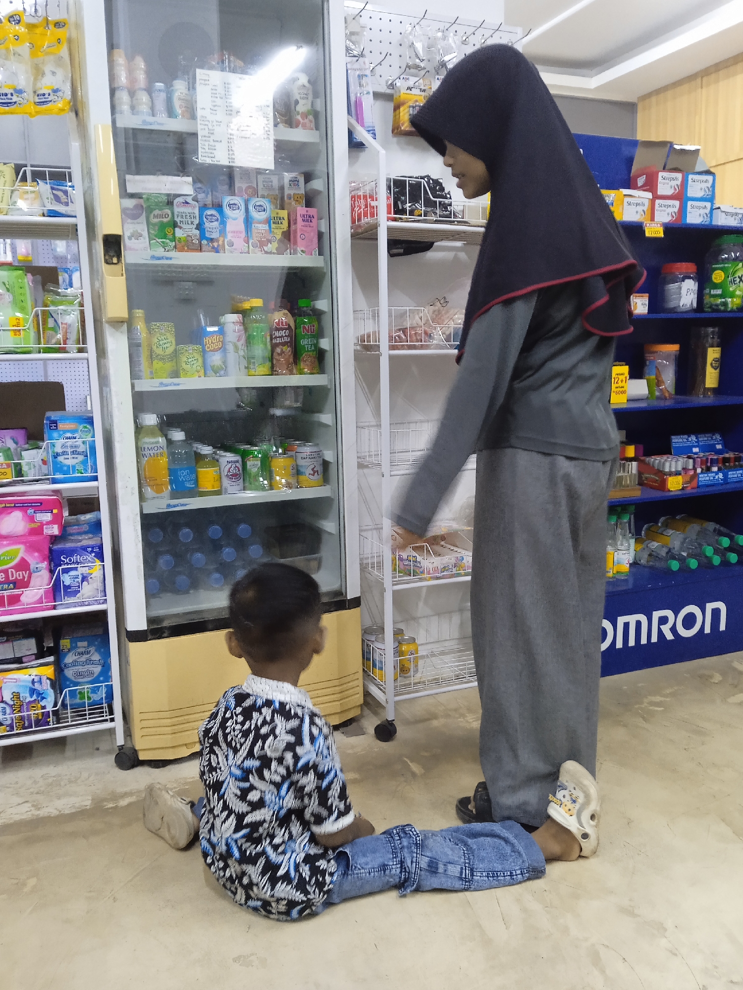 An indoor scene in what appears to be a minimarket or small store in Kebonsari, Indonesia, shows an adult woman and a young child. The woman, wearing a black hijab with red trim, a grey long-sleeved top, and grey pants, stands facing a glass-fronted refrigerator. She wears a black slipper on one foot and a light-colored, possibly Croc-style, sandal on the other. A young child, dressed in a patterned blue, black, and white short-sleeved shirt and blue jeans, sits on the floor in front of the refrigerator, looking into it. The child wears a light-colored slipper on one foot.

The refrigerator is filled with various beverages, including "MILO," "FRESH MILK," "ULTRA MILK," "Hydro COCO," "CHOCO JAZELTRA," "GREEN TEA," "LEMON WATER," and "ion wate," along with products from "Nestle" and "CAP KAKI TIGA." A handwritten price list is taped to the inside of the refrigerator door. To the left of the refrigerator are shelves stocked with household items and personal care products like "CHARM," "Softex," and "Cooling Fresh." To the right, another set of shelves holds items like "Strepsils" and other packaged goods, with visible price signs for "HARGA 17.000" and "12+1 - 6000." A prominent blue unit at the bottom right features the brand name "OMRON." The lighting suggests a well-lit interior, possibly daytime. No specific emotions are discernible from the individuals.