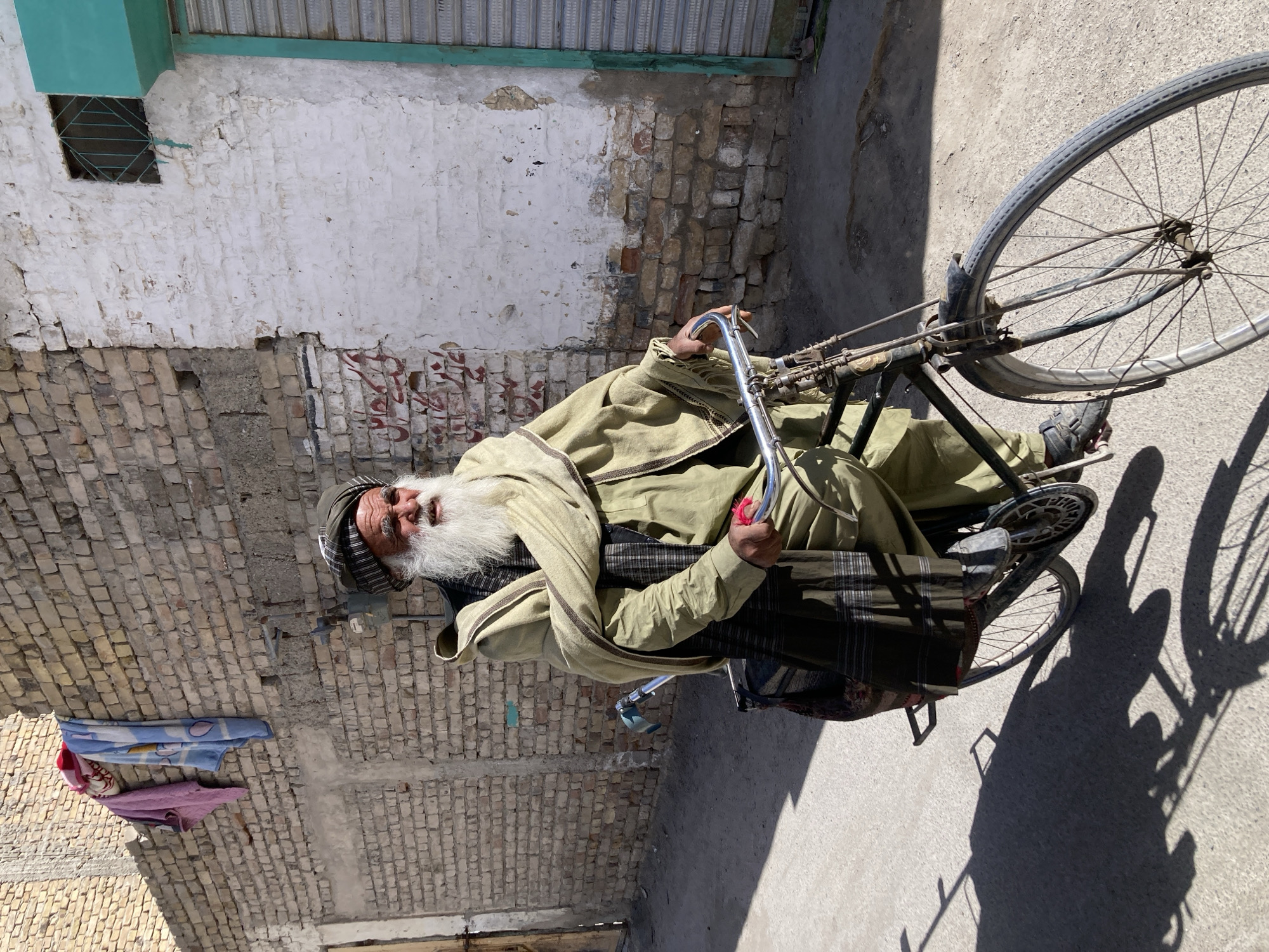 An elderly man with a long white beard and wearing traditional Pakistani clothing sits on a bicycle. He is positioned in front of a building constructed from bricks and painted in varying shades of white and tan. The building shows signs of aging with visible wear and tear. A small window with a metal grate is present on the upper part of the building. To the right of the man and his bicycle is a paved road surface. The image was taken in Quetta City Tehsil, Pakistan.